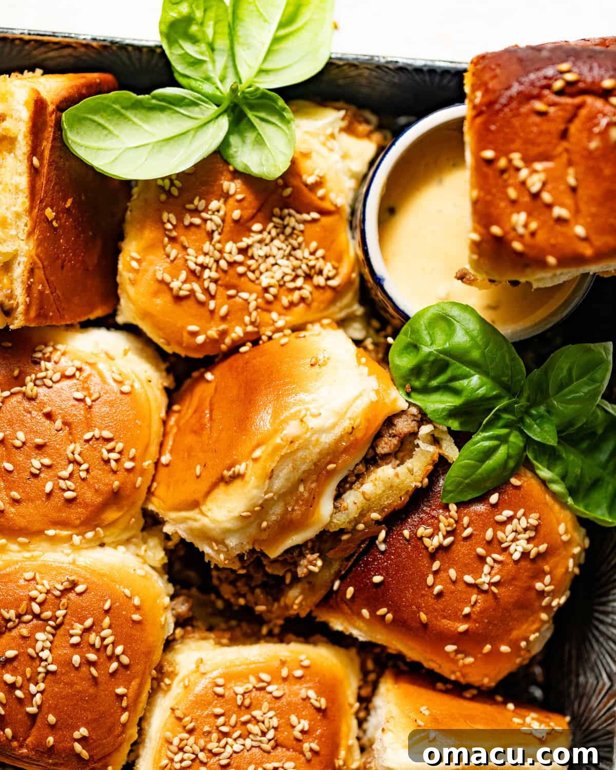 Cheeseburger sliders with Hawaiian rolls served on a wooden board, garnished with fresh basil and a side of burger sauce.
