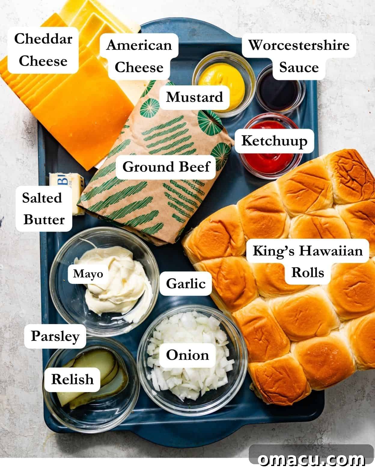 A flat lay image displaying all key ingredients for cheeseburger sliders: ground beef, butter, fresh parsley, minced garlic, ketchup, mustard, relish, King's Hawaiian rolls, American cheese, cheddar cheese, mayonnaise, diced onion, garlic powder, onion powder, and Worcestershire sauce.