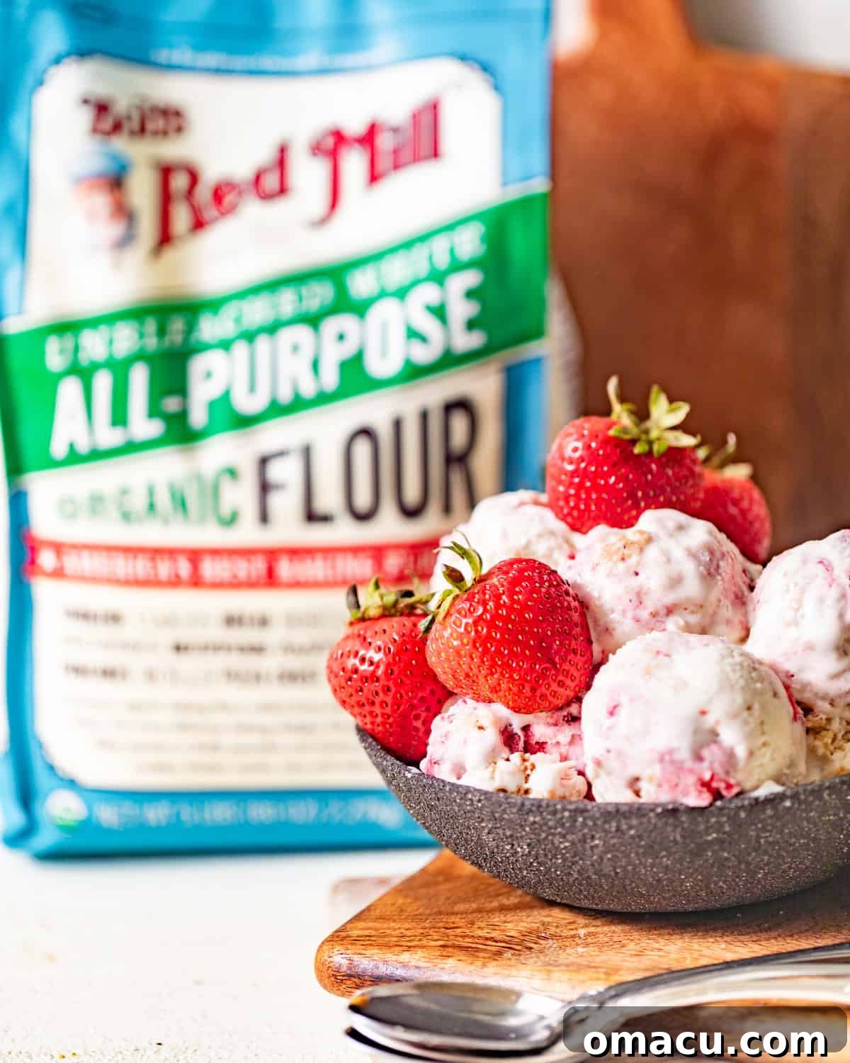 A beautiful bowl of strawberry shortcake ice cream, garnished with fresh whole strawberries, placed in front of a bag of Bob's Red Mill flour, highlighting the quality ingredients used.