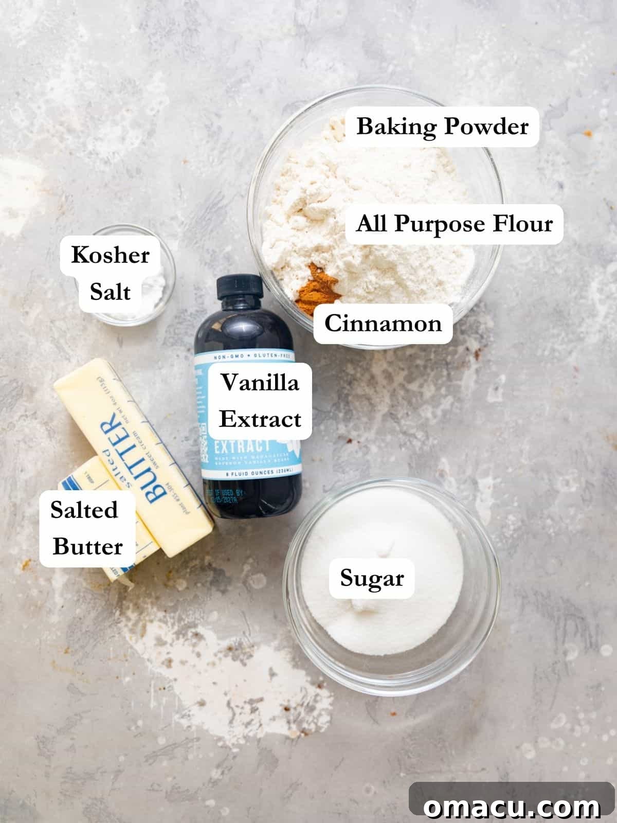 A selection of ingredients for cherry cobbler biscuit topping including flour, vanilla extract, salted butter, sugar, baking powder, salt, and cinnamon, arranged on a rustic wooden surface.