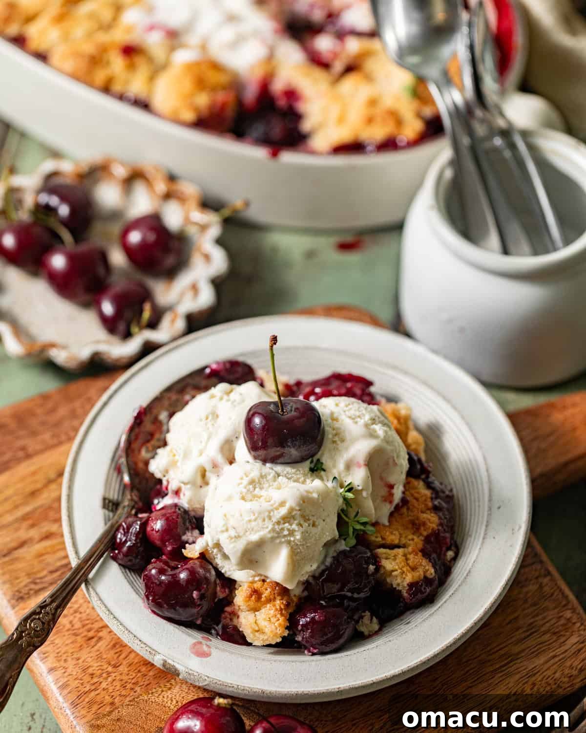 A serving of cherry cobbler in a bowl, topped generously with vanilla ice cream and garnished with fresh cherries, ready to be enjoyed.