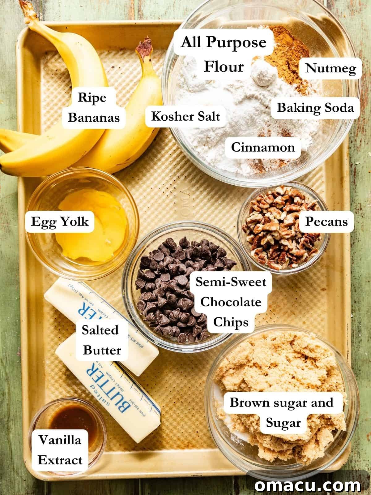 A collection of ingredients laid out on a baking sheet, including ripe bananas, all-purpose flour, cornstarch, baking soda, spices like cinnamon and nutmeg, brown sugar, granulated sugar, vanilla extract, melted butter, semi-sweet chocolate chips, and chopped pecans.