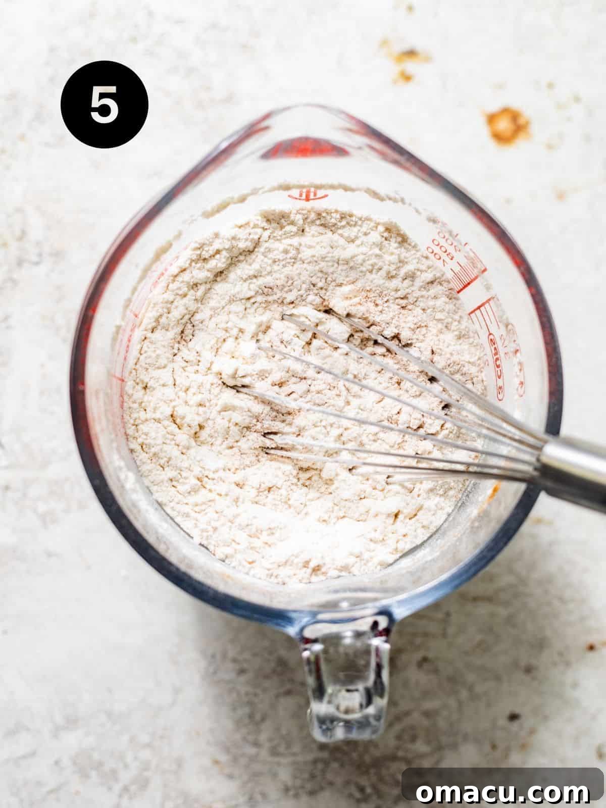 Cake flour, pumpkin pie spice, baking powder, and kosher salt are meticulously mixed together in a bowl, representing the combined dry ingredients for the donut batter.