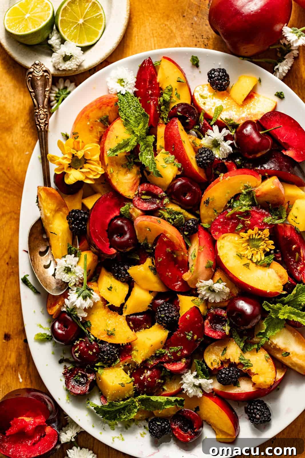 A vibrant stone fruit salad elegantly arranged in a white serving bowl, garnished with delicate edible flowers and fresh lime slices, accompanied by a polished serving spoon, ready to be enjoyed.