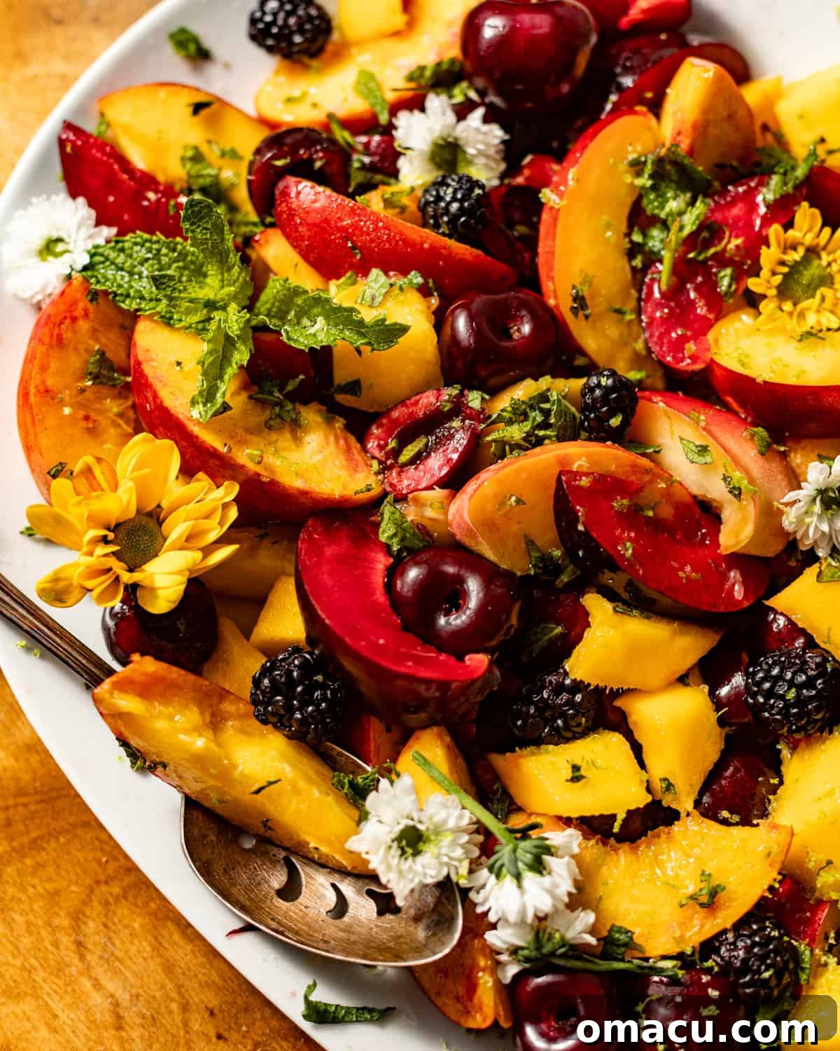 A close-up photograph of a vibrant and appetizing stone fruit salad, emphasizing the juicy textures and rich colors of the peaches, plums, cherries, mangoes, and blackberries, beautifully garnished with delicate fresh mint leaves.