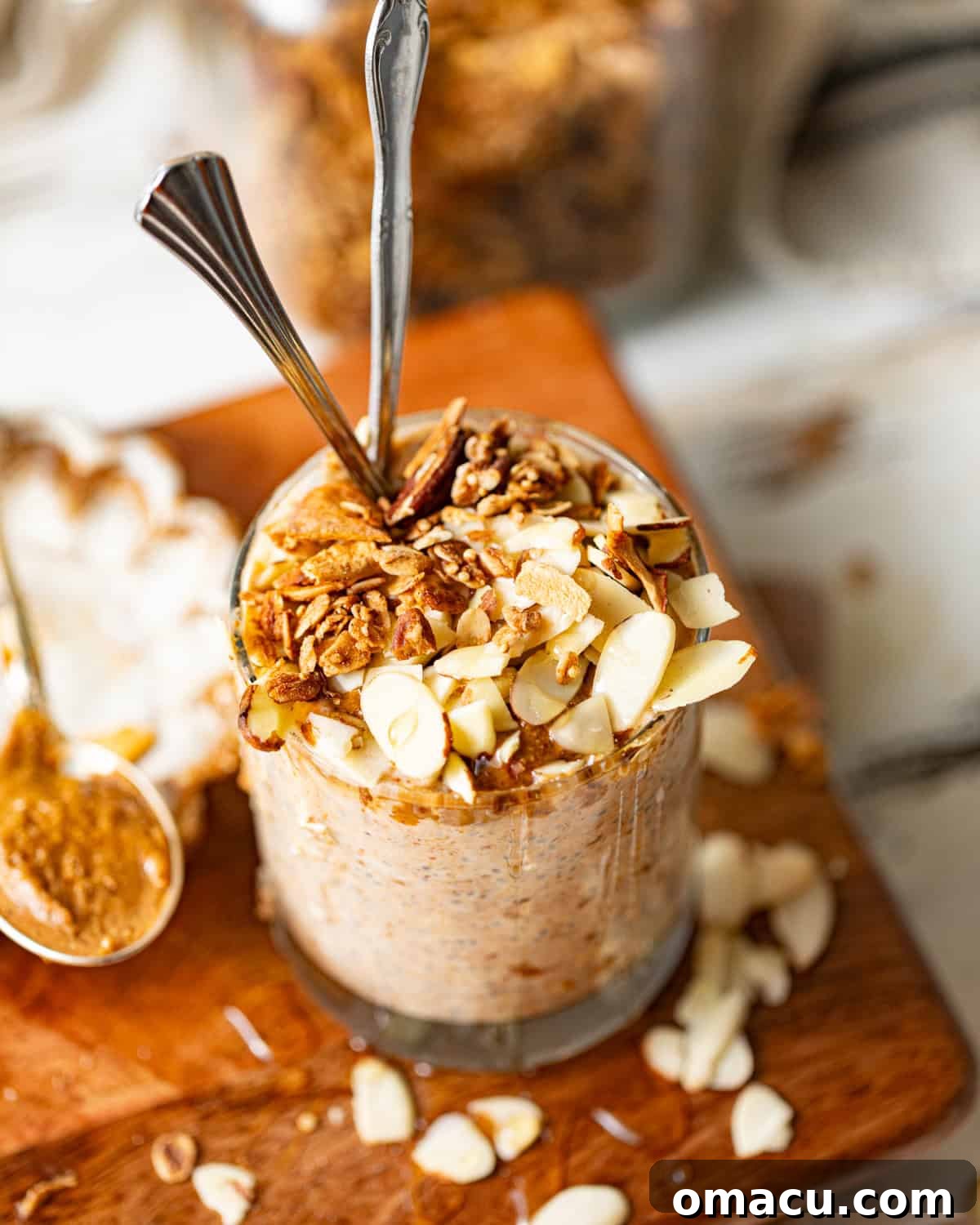 A serving of almond butter overnight oats in a stylish cup, generously drizzled with extra almond butter and topped with crunchy sliced almonds, presented on a rustic wooden board.