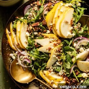 Arugula Pear Salad in a gold dish with spoons and sliced pears next to the dish.