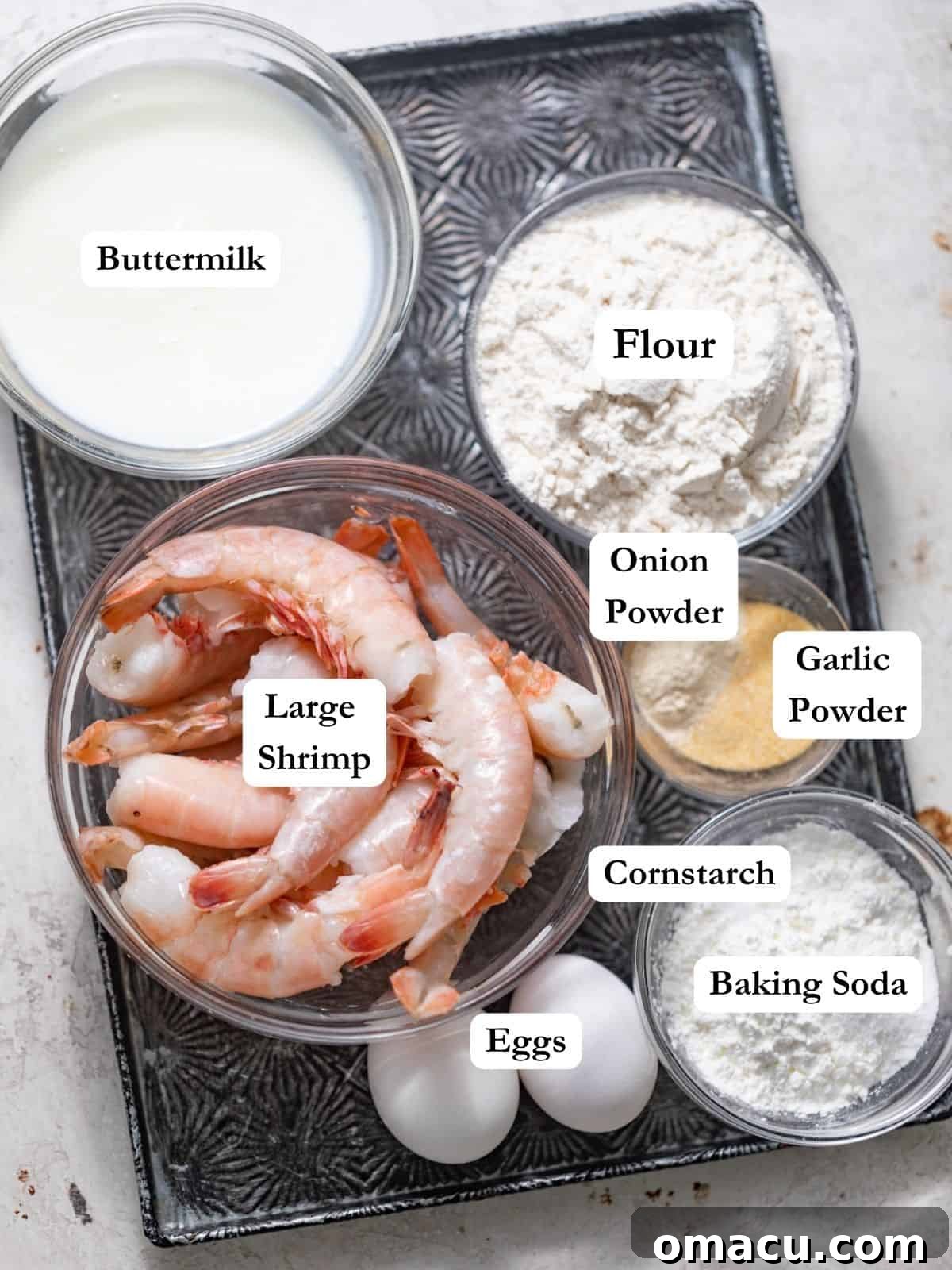 An assortment of fresh ingredients laid out on a tray, including large raw shrimp, all-purpose flour, cornstarch, buttermilk, garlic powder, onion powder, baking soda, kosher salt, and eggs, all essential for making crispy Bang Bang Shrimp.