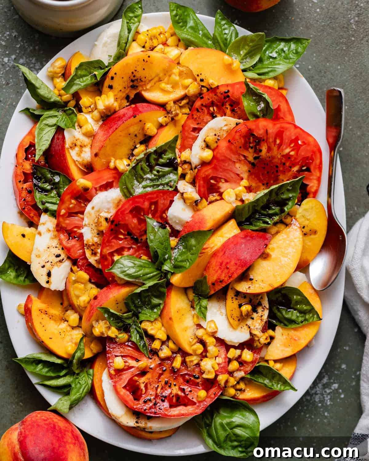 A vibrant Peach Caprese Salad on a white plate, artfully arranged with slices of fresh peaches, heirloom tomatoes, fresh mozzarella, basil leaves, and golden sautéed corn. It's garnished with a rich balsamic glaze and a sprinkle of flaky sea salt, highlighting its fresh, gourmet appeal.