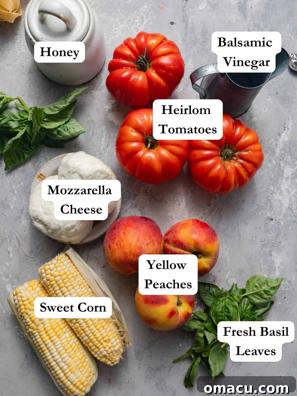 Arrangement of fresh ingredients for Peach Caprese Salad, including whole and sliced peaches, vibrant heirloom tomatoes, fresh basil sprigs, yellow corn kernels, a ball of fresh mozzarella, bottles of balsamic vinegar and honey, and a pinch of flaky sea salt.