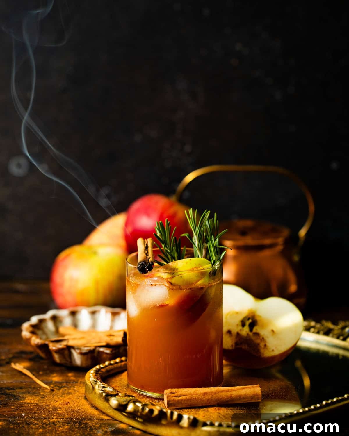 An elegant apple cider cocktail in a glass, placed on a golden tray and garnished with rosemary and fresh apple slices.
