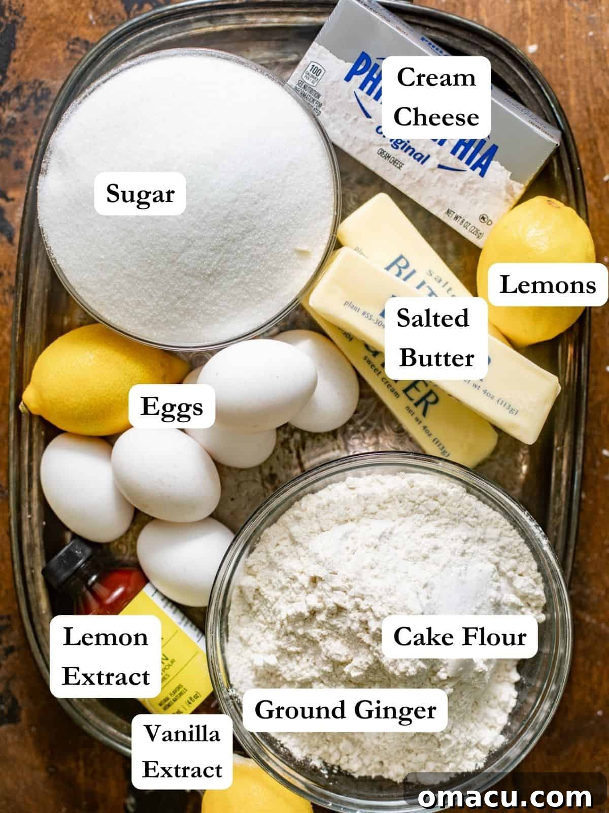 All the essential ingredients for lemon pound cake laid out: sugar, butter, cream cheese, lemon zest, cake flour, ginger, eggs, and kosher salt.