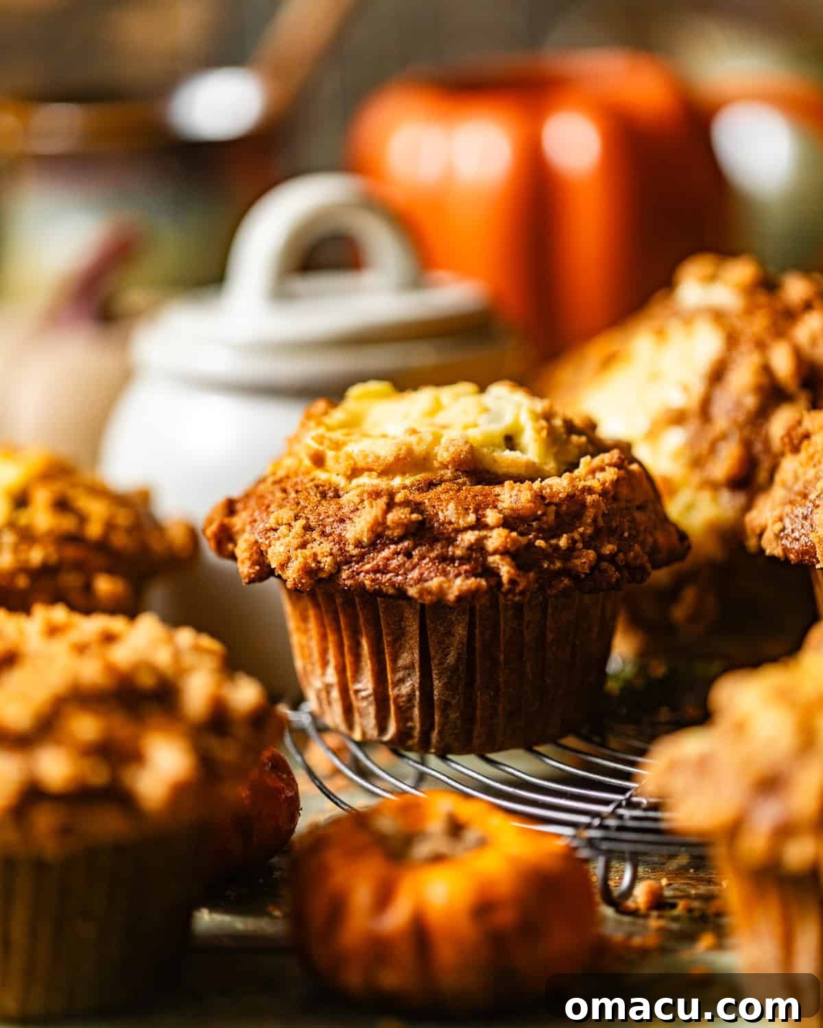 A close-up of freshly baked pumpkin cream cheese muffins cooling on a wire rack, with a decorative jar and a festive pumpkin mug in the background, hinting at cozy fall moments.