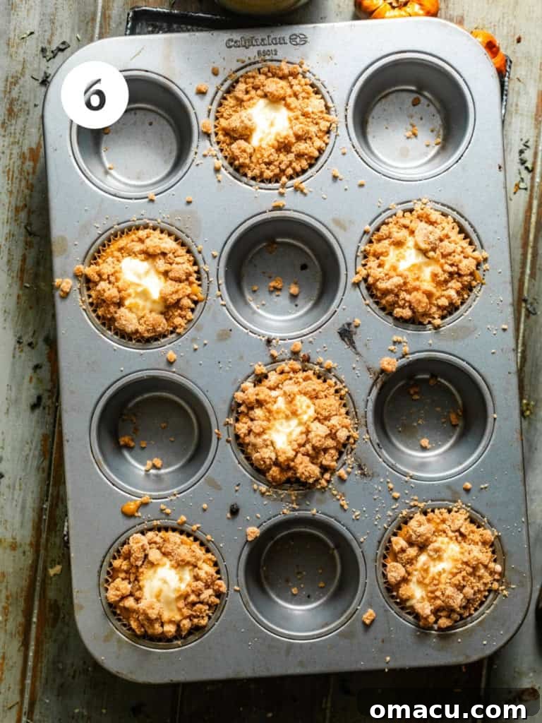 A muffin pan filled with pumpkin cream cheese muffins, each generously topped with a dollop of creamy filling and a sprinkle of cinnamon streusel, perfectly arranged and ready for the oven.