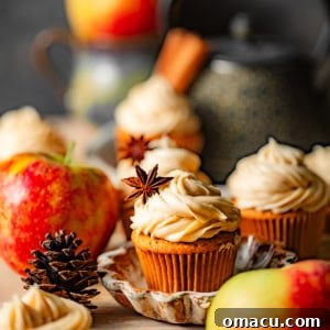 apple cider cupcakes filled with apple butter topped with cinnamon cream cheese frosting surrounded by apples on a cutting board.