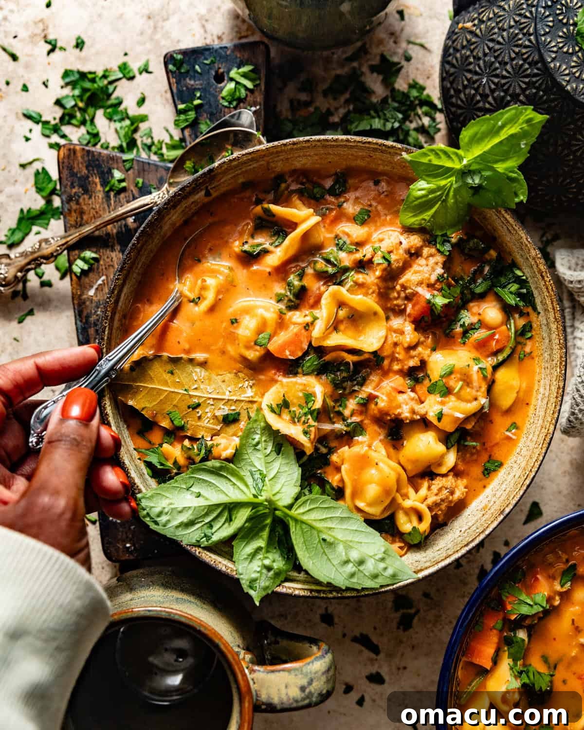 A close-up shot of a hand holding a spoon, scooping up a portion of the rich and creamy Tuscan soup, clearly showing sausage pieces, spinach, and tortellini.