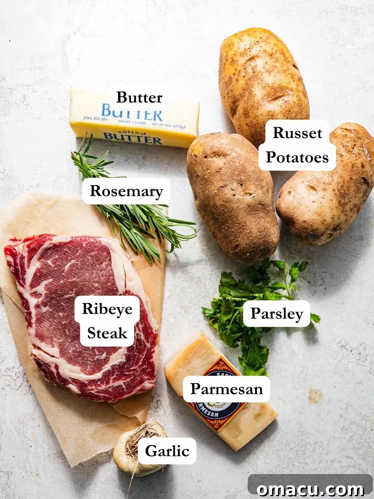 Ingredients to make steak frites - ribeye steak, rosemary, butter, potatoes, parmesan, parsley, and garlic laid out on a table.