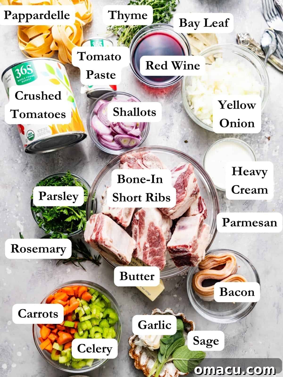 Various fresh ingredients laid out on a white surface, including beef short ribs, bacon, carrots, celery, onions, garlic, fresh herbs, tomato paste, and a bottle of red wine, all ready for making bolognese.