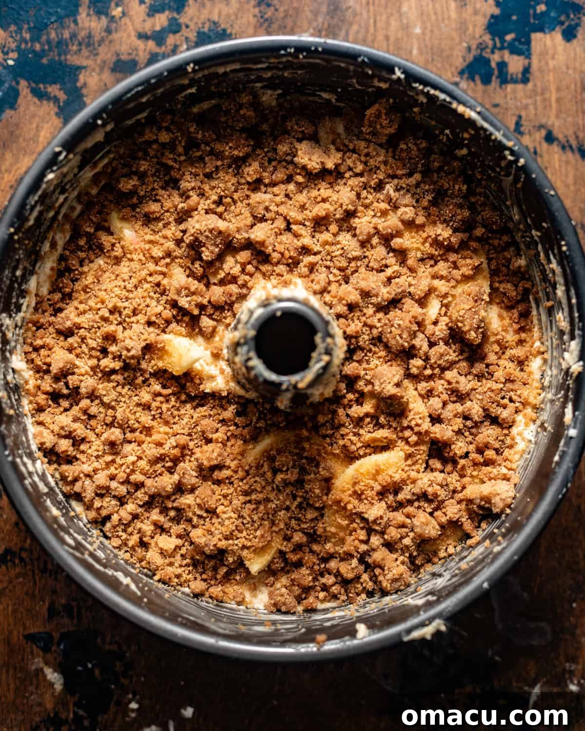 A salted caramel apple crumb cake in a tube pan, generously covered with a golden-brown crumb topping, ready for baking.