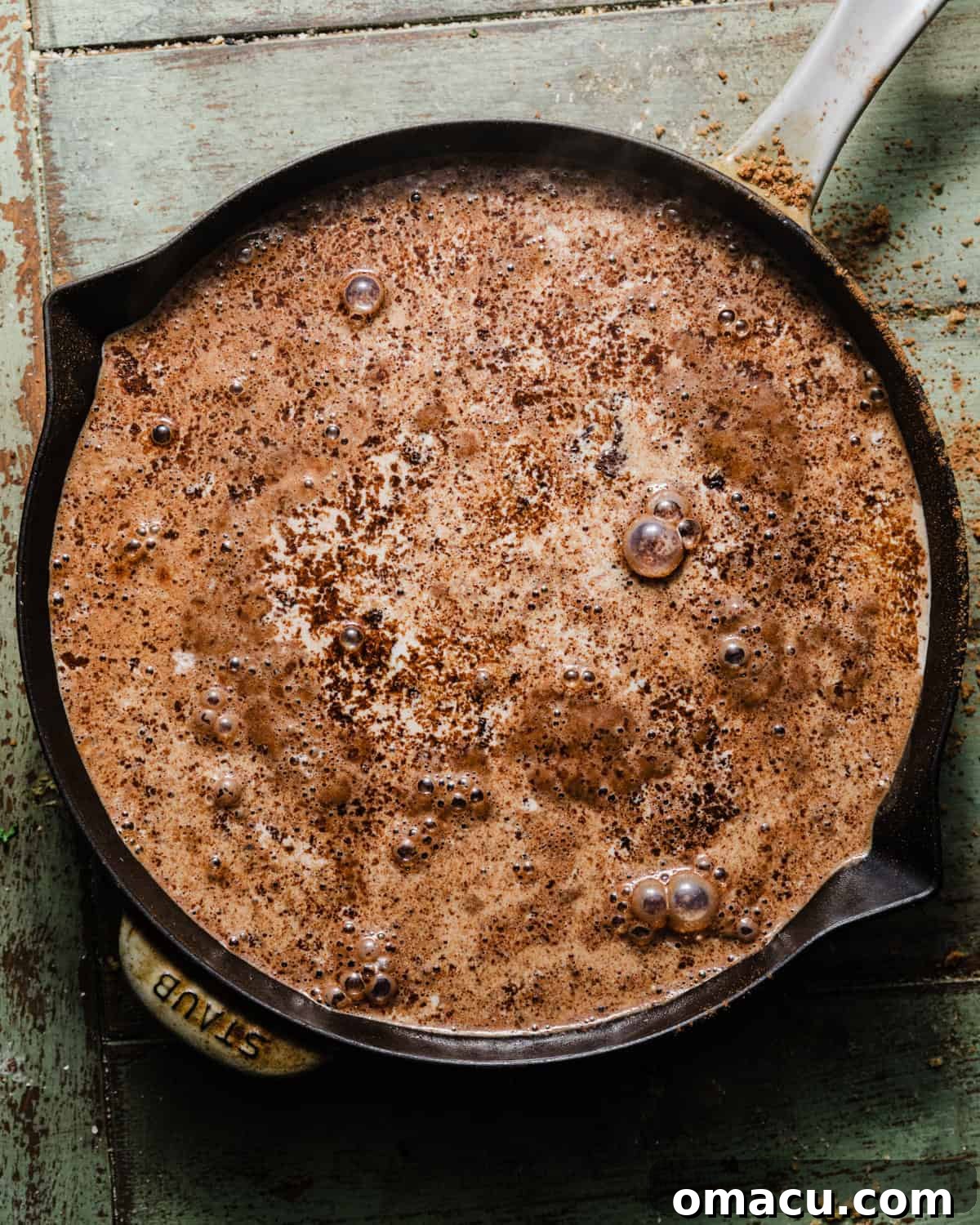 Boiling hot milk and coffee mixture being carefully poured over the chocolate pudding cake batter, which is already topped with brown sugar and cocoa powder, in a skillet.
