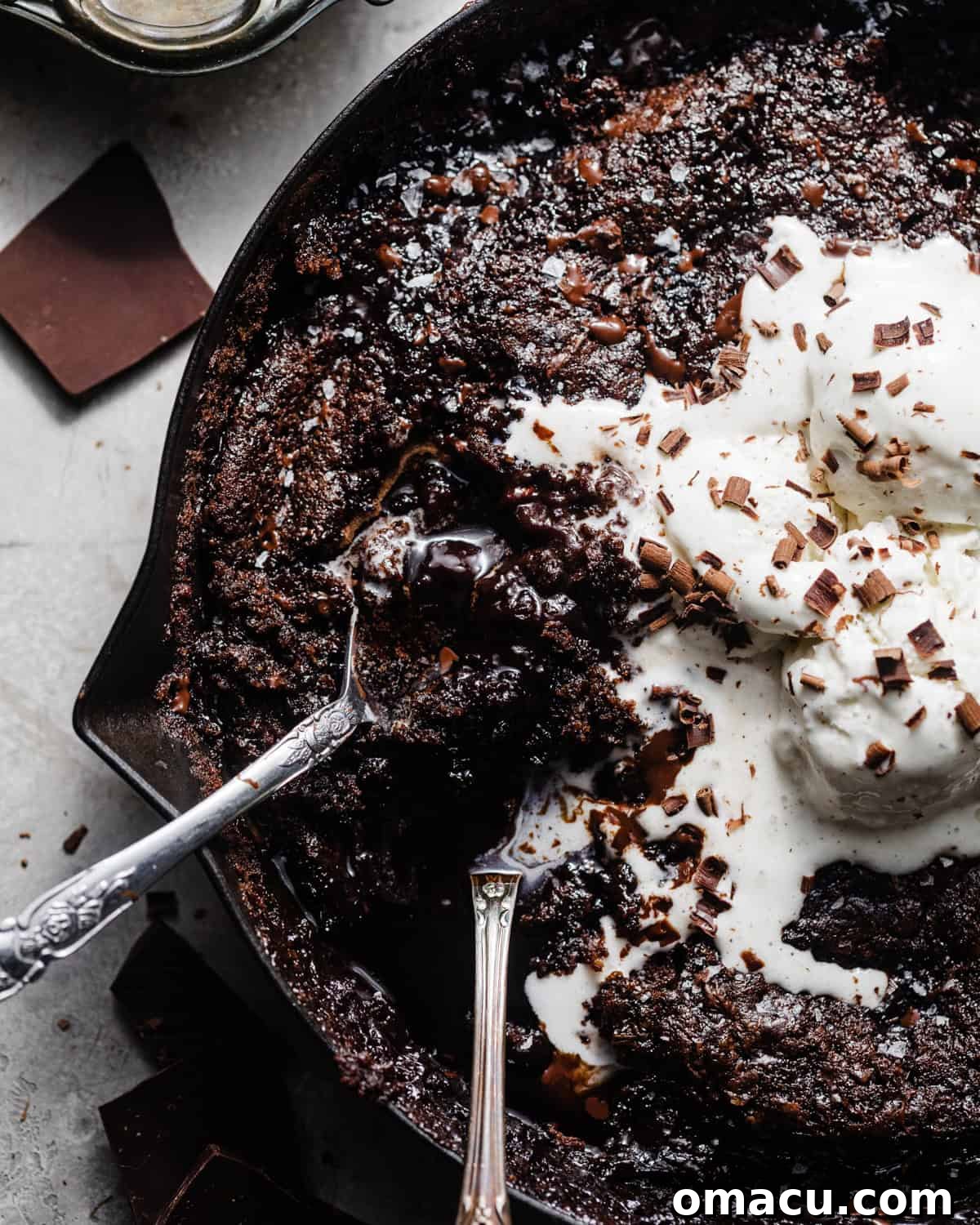 A close-up view of a chocolate pudding cake in a skillet, adorned with melting ice cream. Two spoons are poised to scoop into the warm, gooey dessert, highlighting its irresistible texture.