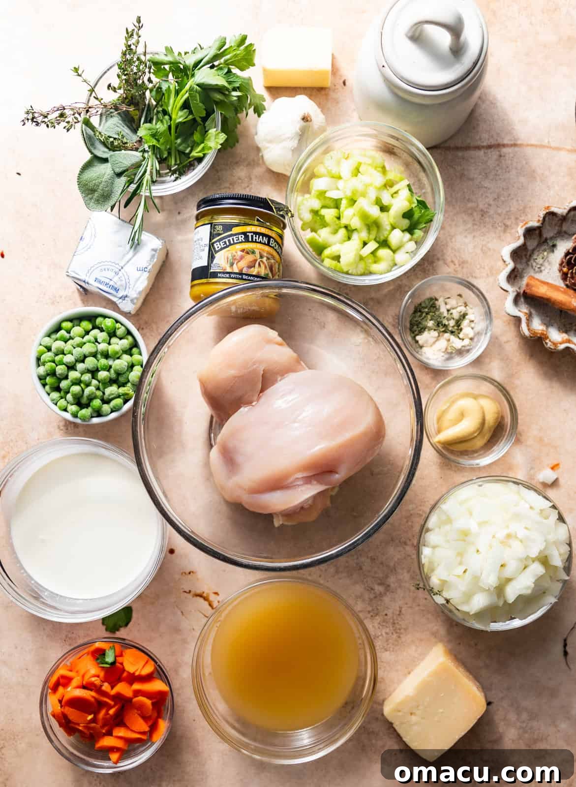 Arrangement of fresh ingredients for chicken pot pie including chicken breasts, carrots, celery, onions, frozen peas, fresh herbs, heavy cream, garlic, butter, chicken broth, chicken bouillon paste, parmesan cheese, and various seasonings.