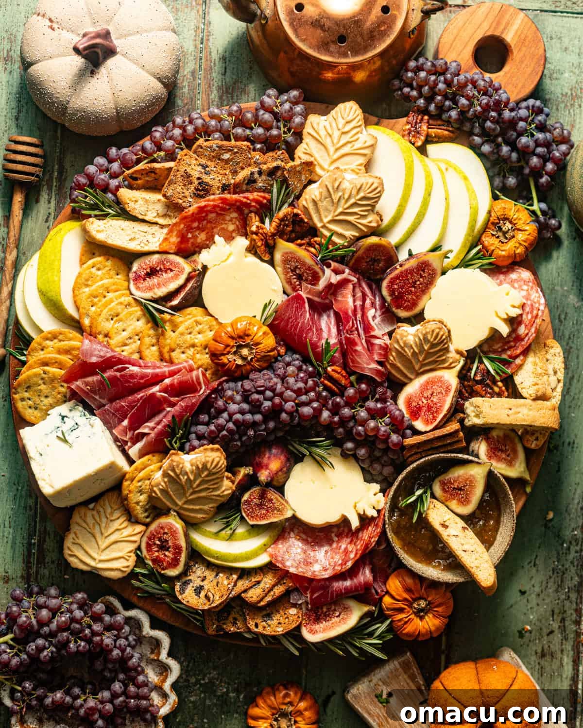 A festive Fall Charcuterie Board arranged on a green wooden surface, surrounded by mini pumpkins and a coffee pot, highlighting autumnal colors and treats.