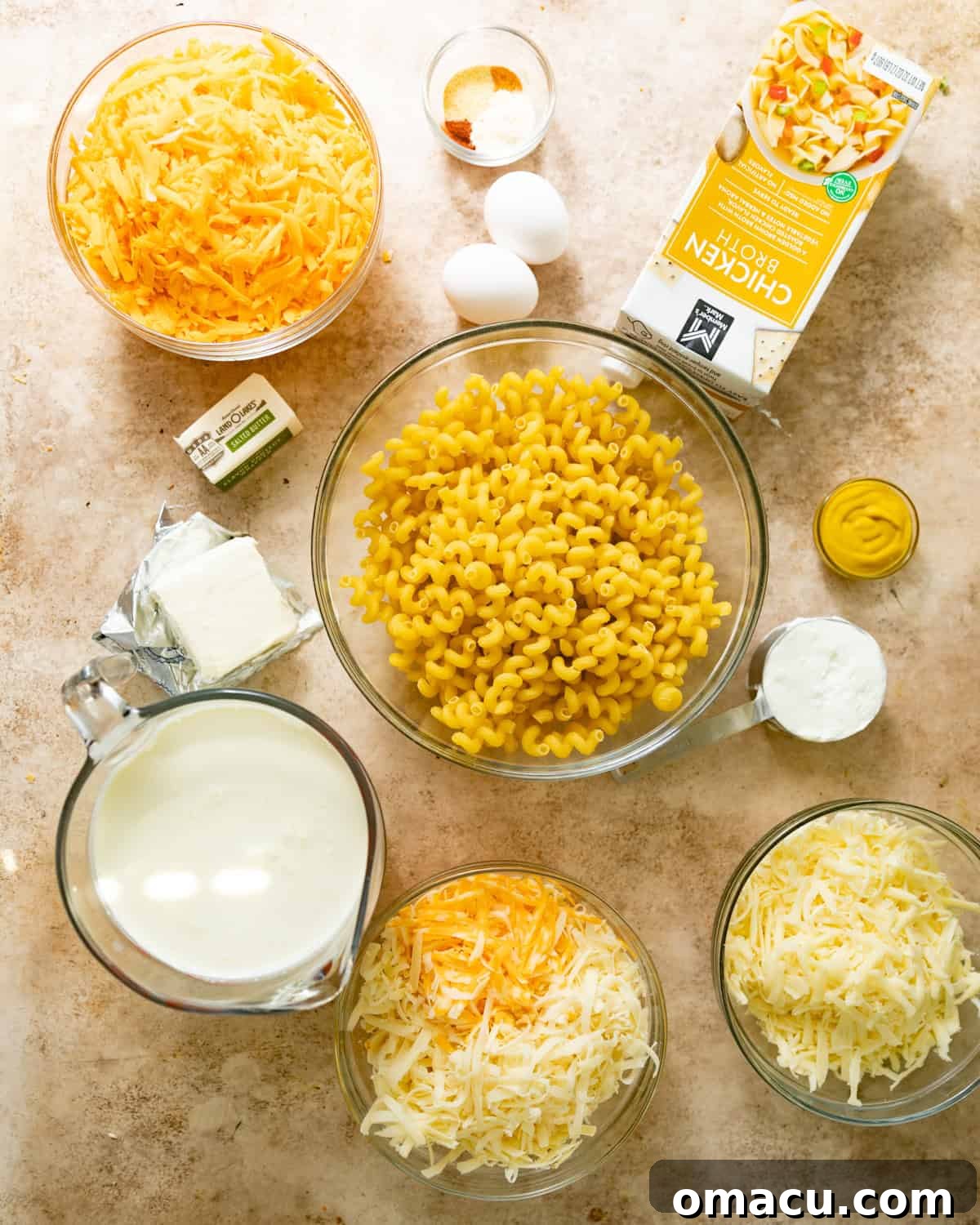 Ingredients laid out for making mac and cheese: cavatappi pasta, chicken broth, various cheeses including sharp cheddar, gruyere, mozzarella, colby jack, evaporated milk, heavy cream, cream cheese, butter, eggs, cornstarch, seasonings, and breadcrumbs.