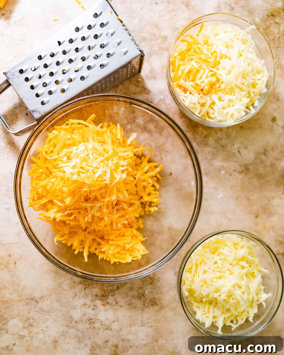 Bowls of various shredded cheeses, including mozzarella, cheddar, Gruyère, and Colby Jack, prepared on a light surface.