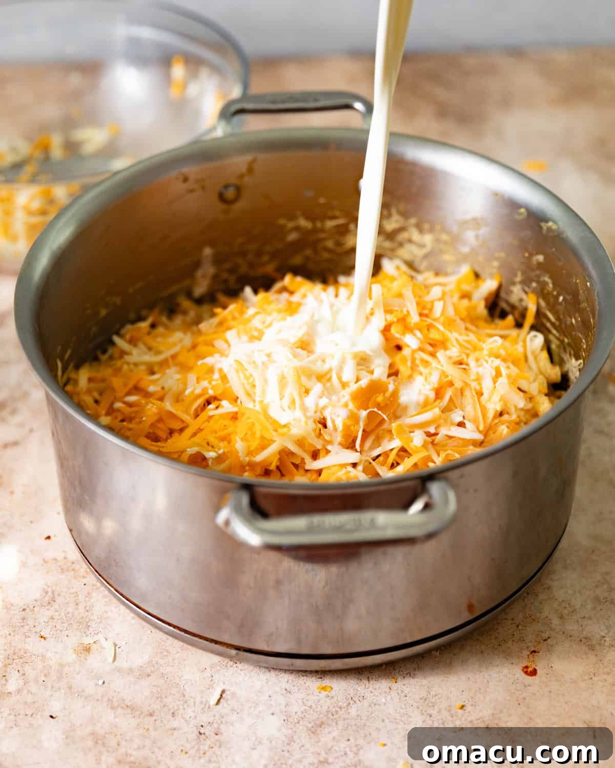 A large pot on a stove with creamy cheese sauce, and a hand pouring milk into the pot, surrounded by melted cheeses.