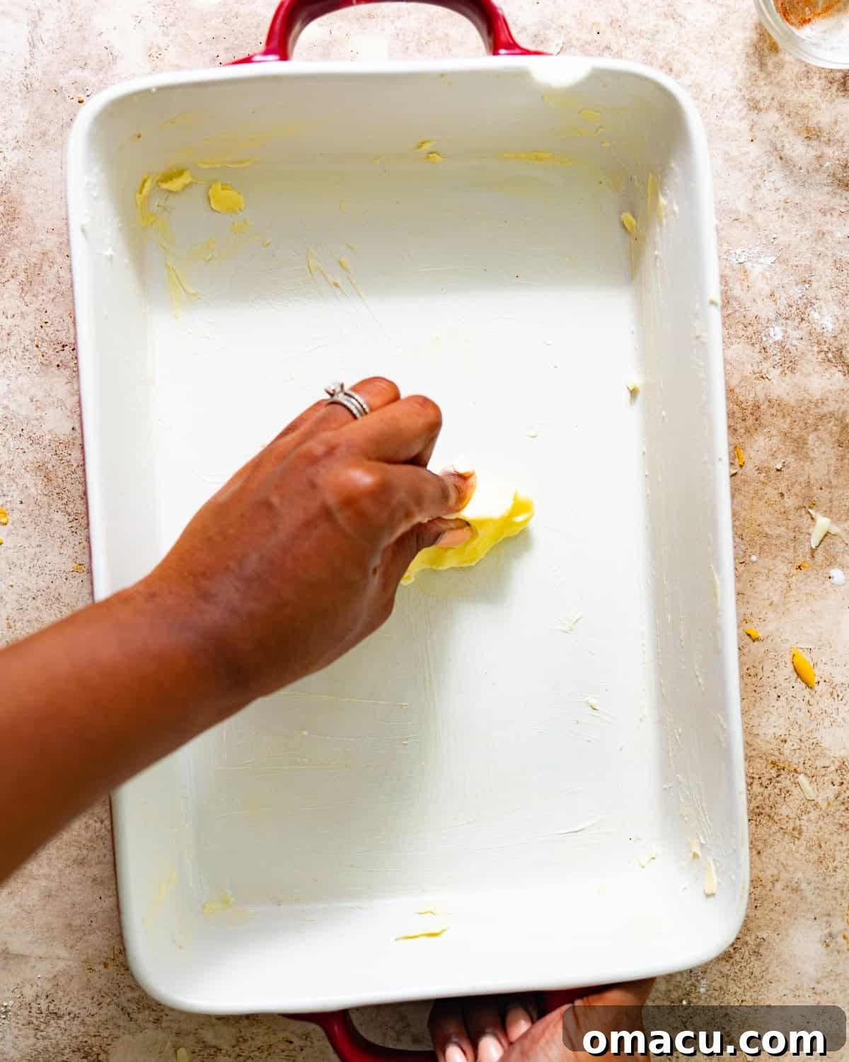 A hand is shown expertly smearing softened butter across the bottom and sides of a clear glass baking dish, preparing it for the mac and cheese.