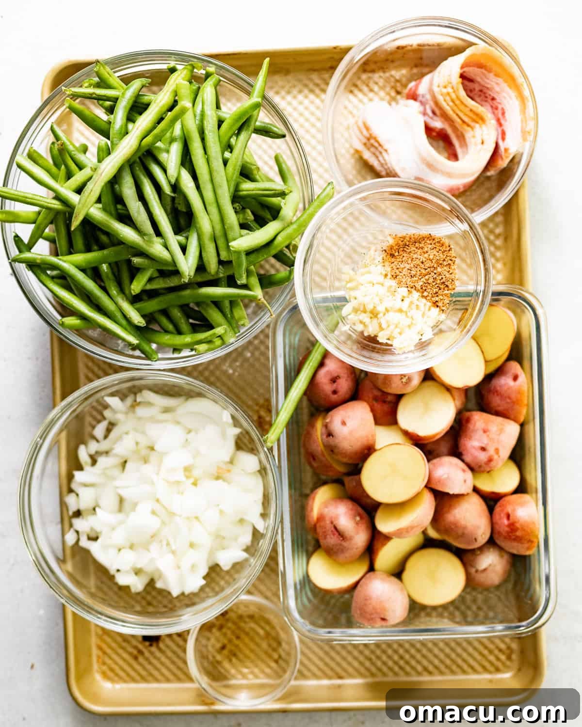 A vibrant display of fresh ingredients: green beans, crispy bacon, yellow onion, garlic cloves, chicken broth, apple cider vinegar, Worcestershire sauce, and a mix of seasonings like garlic powder, onion powder, and cajun seasoning, all neatly arranged for a green beans and potatoes recipe.