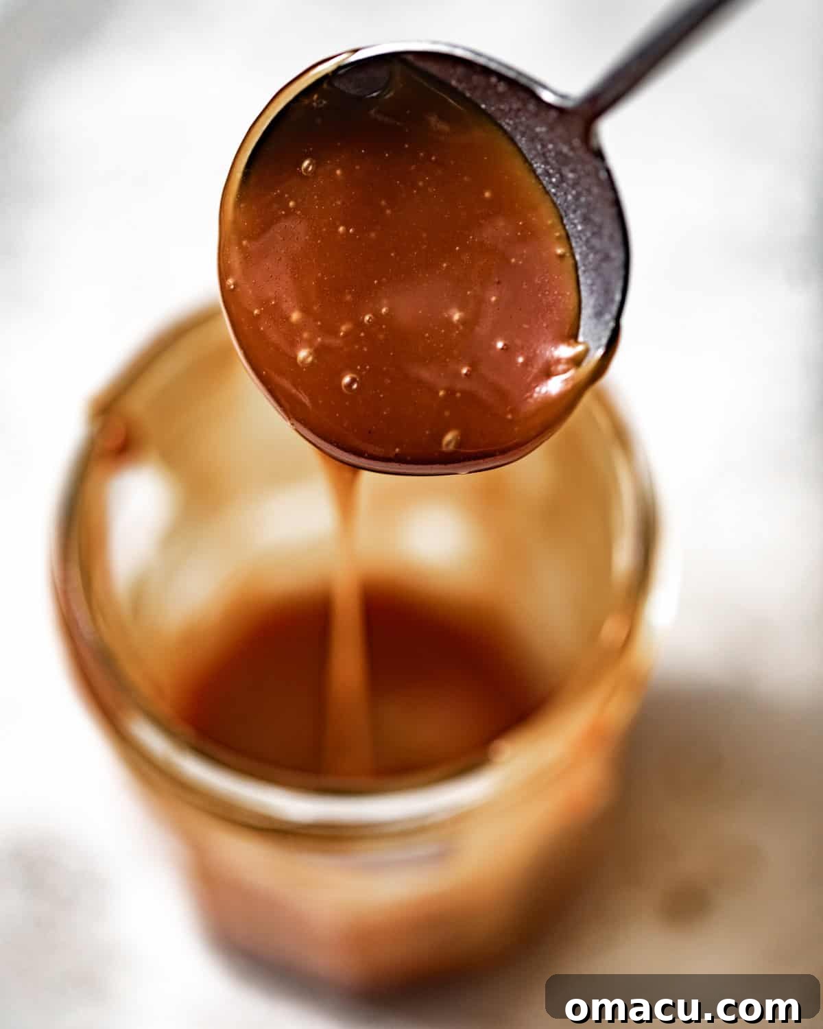 Homemade salted caramel sauce in a clear glass jar with a spoon, showing its rich, amber color and smooth texture.