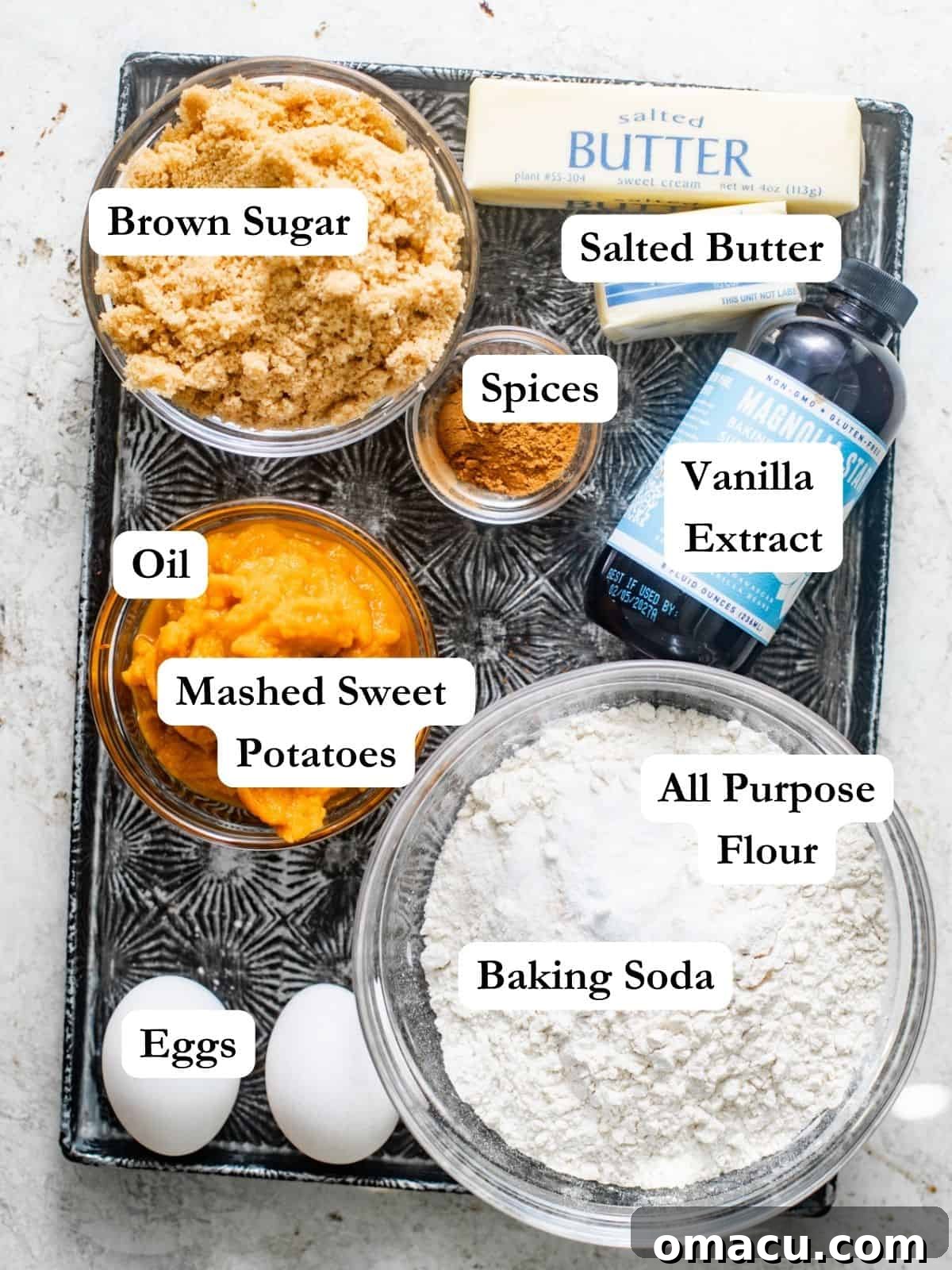 A flat lay photograph showcasing all the ingredients required to make sweet potato bread on a rustic baking sheet, including whole sweet potatoes, butter, oil, various spices, vanilla extract, flour, baking soda, kosher salt, eggs, and brown sugar, ready for preparation.