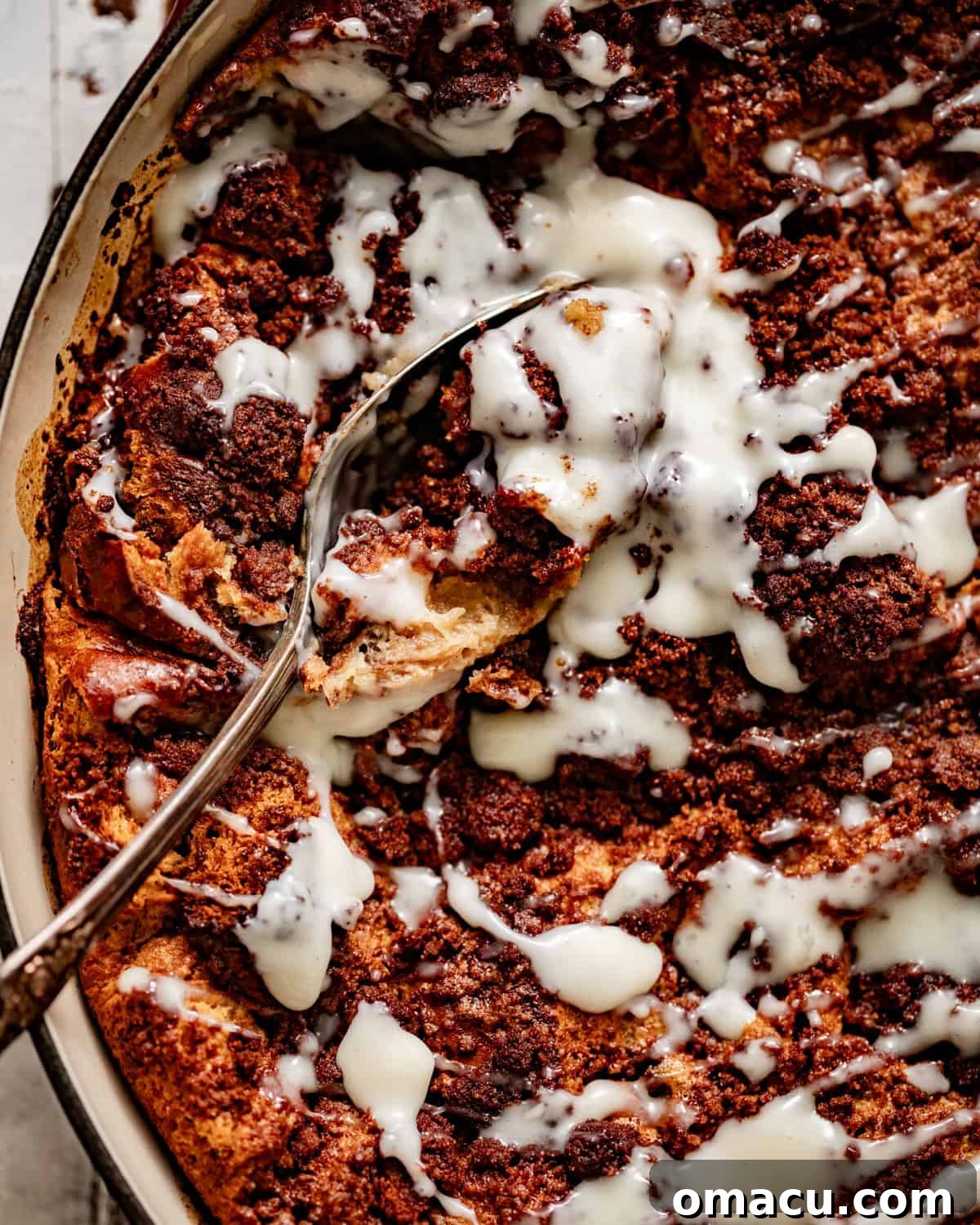 A baking pan filled with golden-brown bread pudding, drizzled with vanilla sauce, with a serving spoon resting inside.