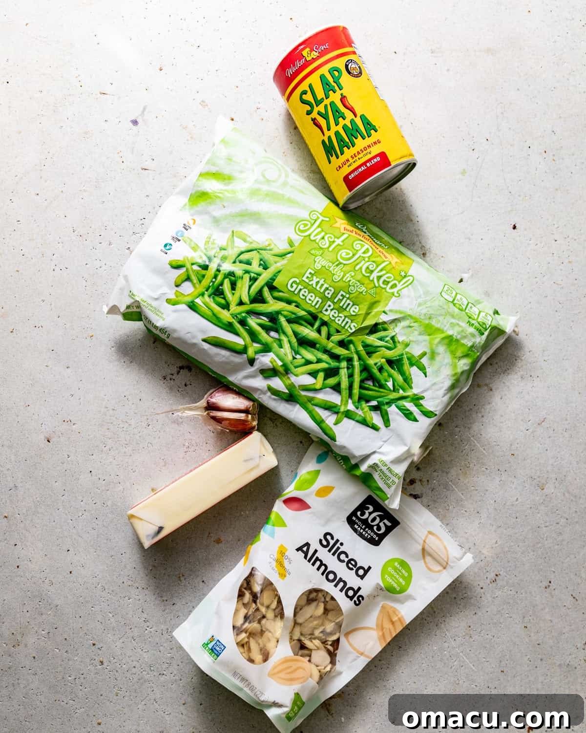 Ingredients to make roasted green beans on a white surface - frozen green beans, olive oil, cajun seasoning, salt, pepper, butter, and garlic.