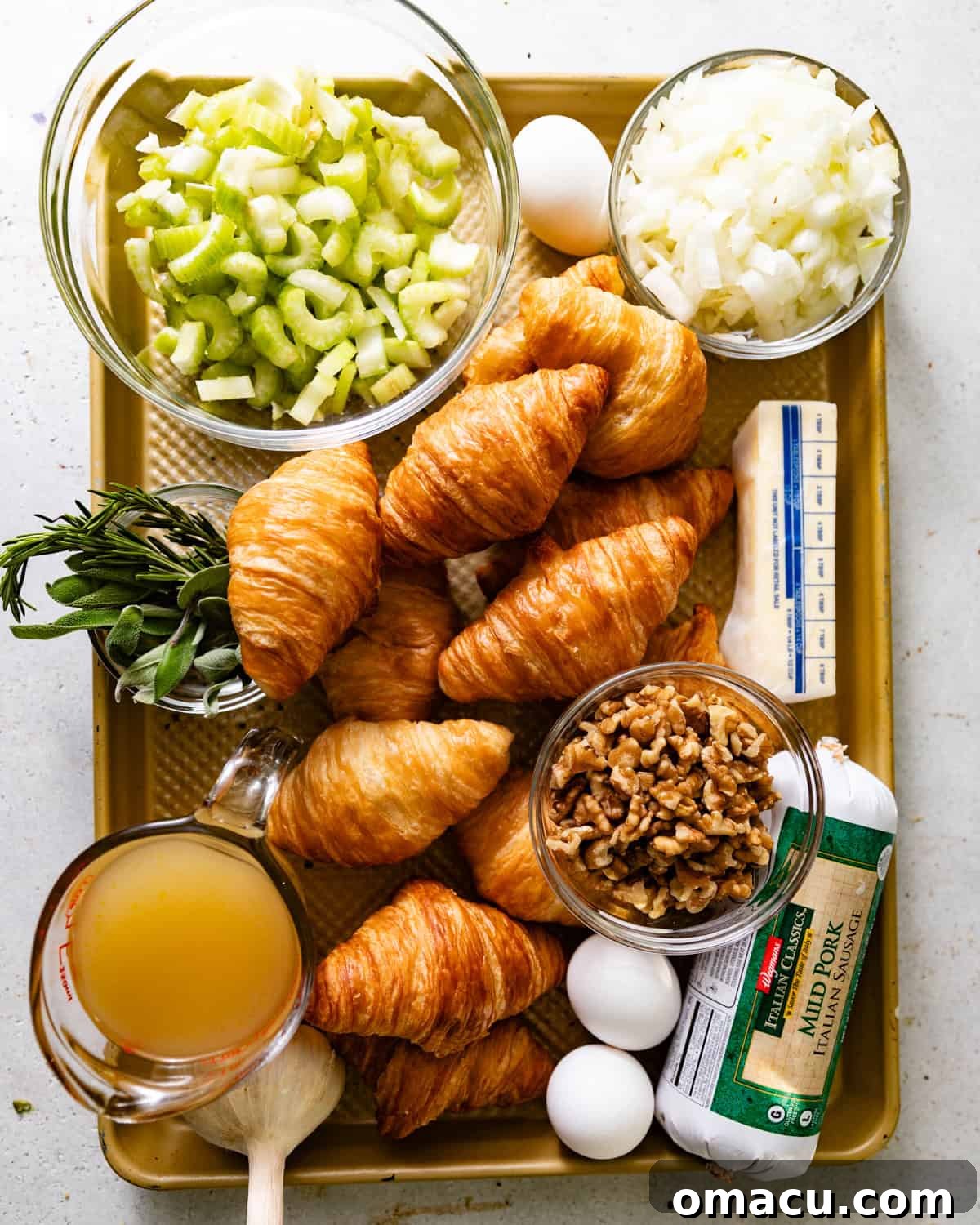 A spread of fresh ingredients for croissant stuffing: whole croissants, diced onion and celery, garlic cloves, chicken broth, eggs, butter, raw breakfast sausage, and a bundle of fresh herbs including thyme, rosemary, sage, and parsley.