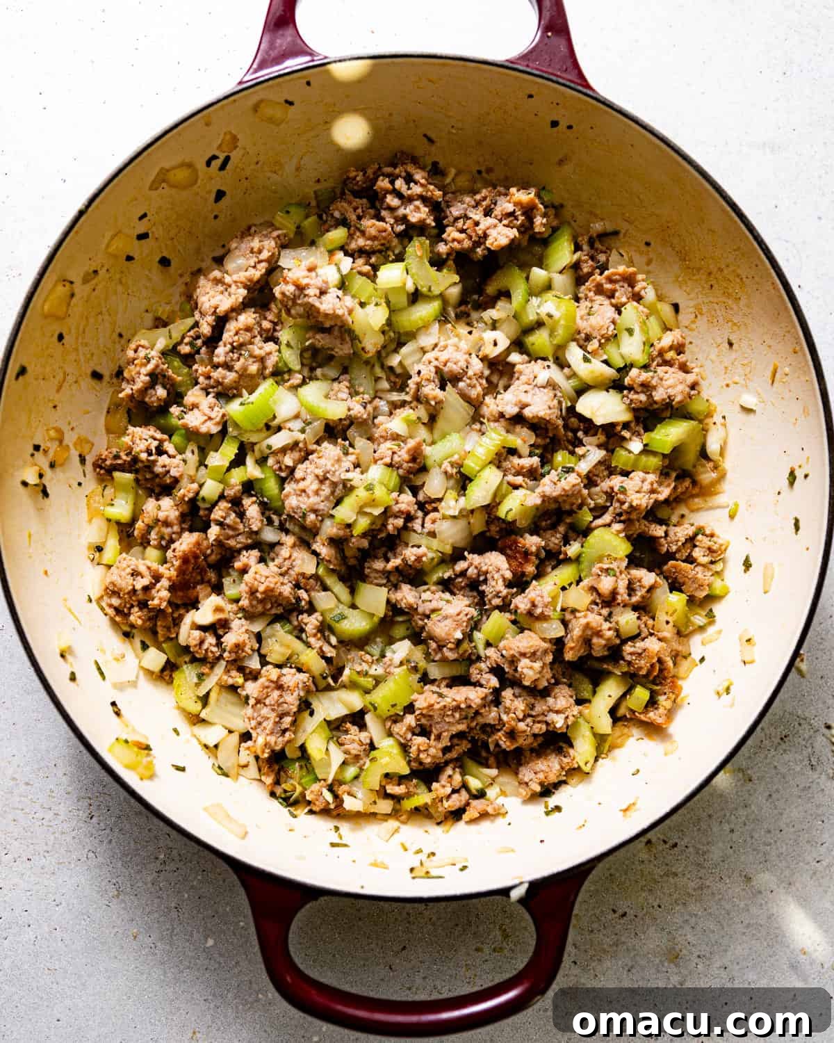 A savory mixture of browned breakfast sausage, softened celery, onions, and garlic, infused with fresh herbs, cooking in a large skillet.