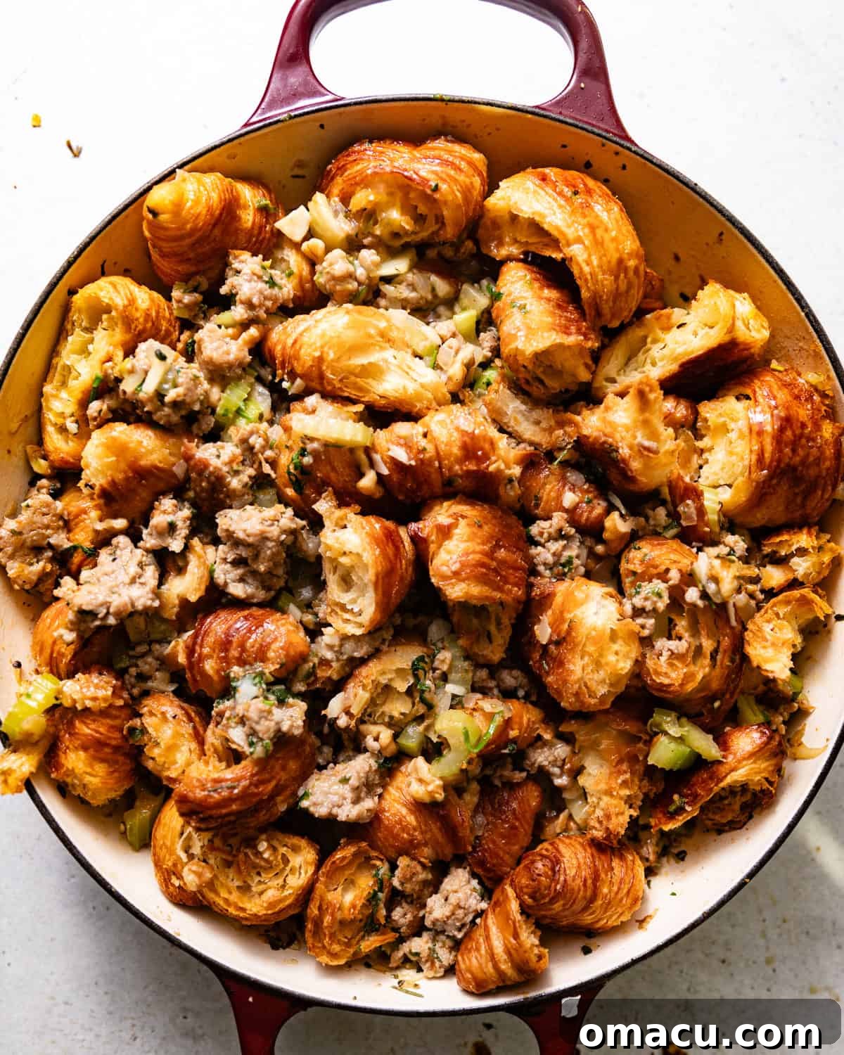 Assembled croissant stuffing, a mix of toasted croissant pieces, savory sausage, and vegetables, spread evenly in a rectangular baking pan, ready for the oven.