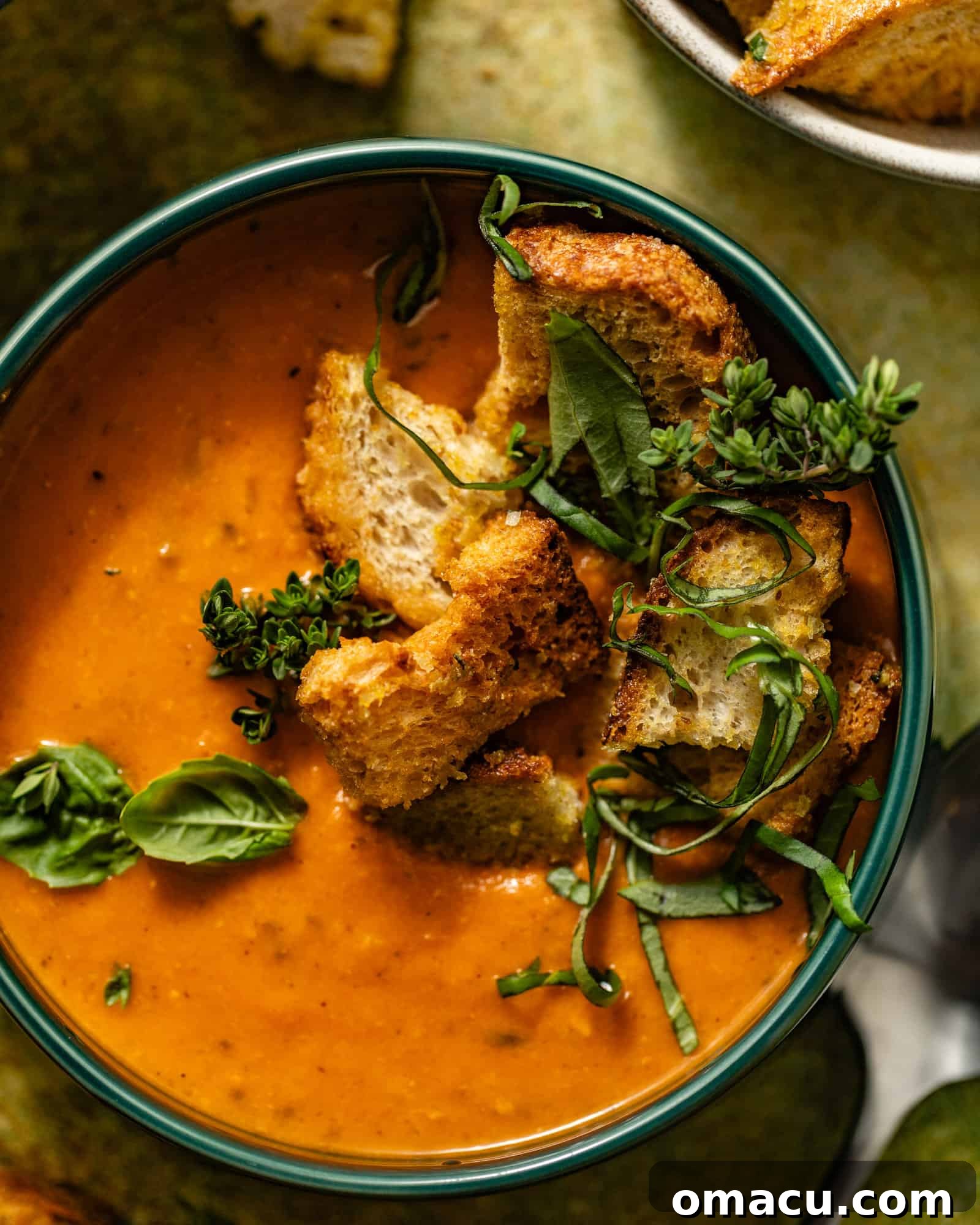 A bowl of creamy heirloom tomato soup, garnished with fresh herbs, crispy garlic croutons, and spoons resting on the side.