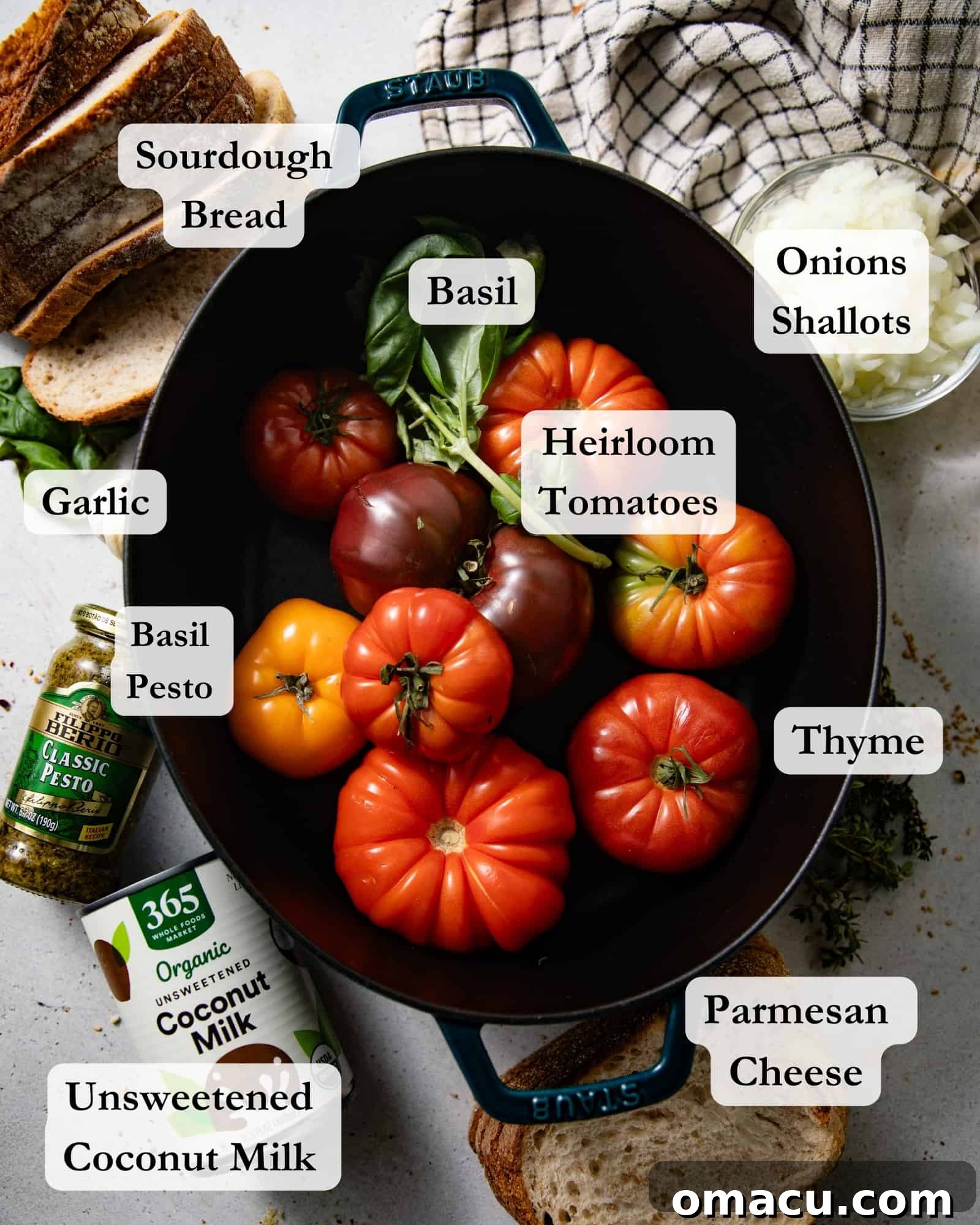 Ingredients for homemade heirloom tomato soup arranged in bowls on a white table: fresh heirloom tomatoes, thyme, basil, garlic, pesto, coconut milk, bread for croutons, olive oil, onions, and shallots.