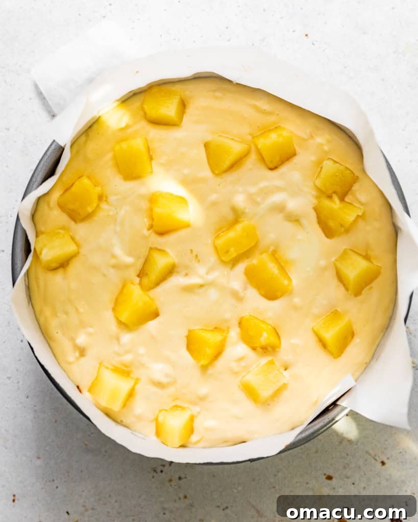 Pineapple cheesecake batter being carefully poured into the prepared Biscoff cookie crust in a springform pan.