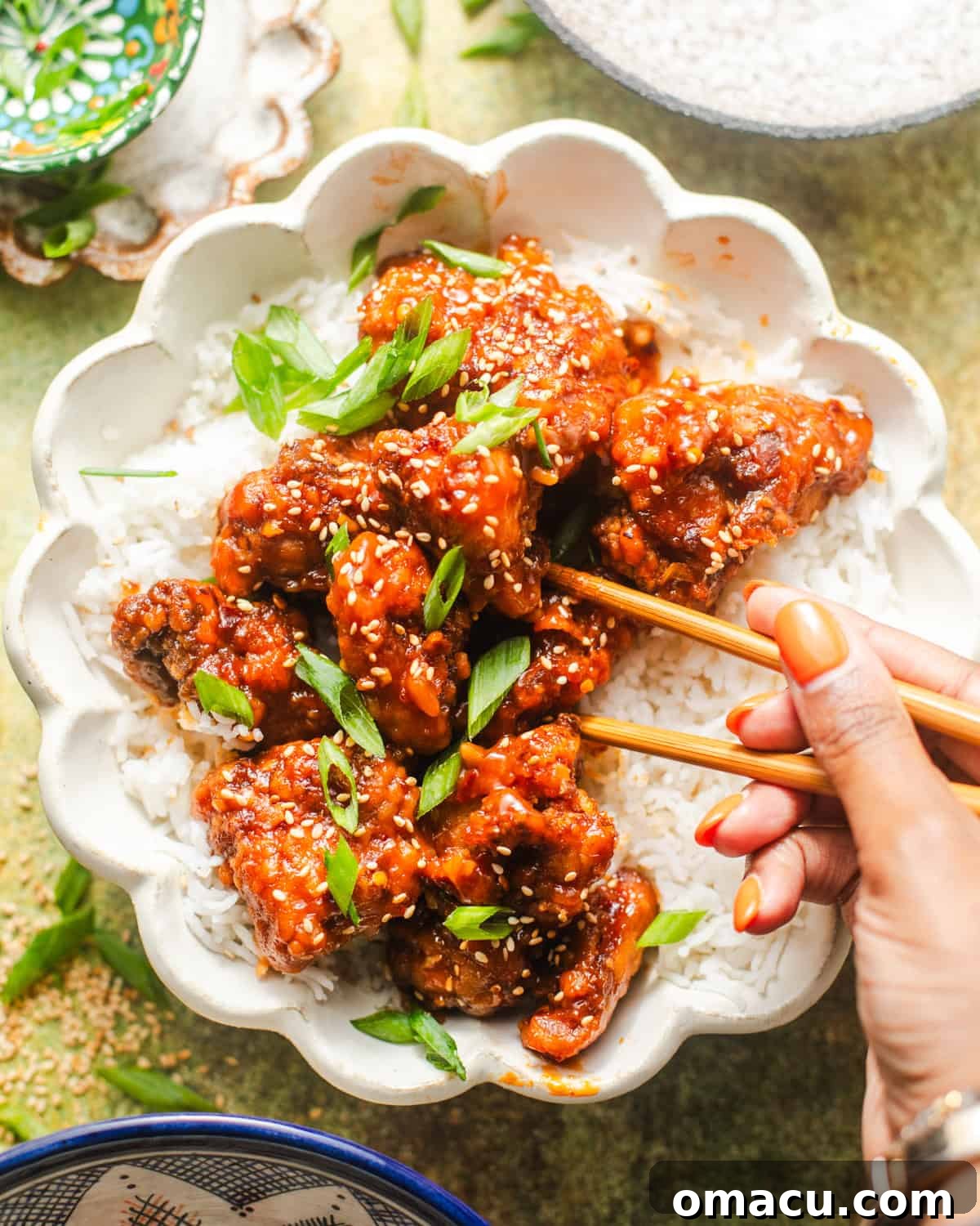 A close-up of crispy orange chicken and rice in a bowl, with a hand holding chopsticks picking up a piece of chicken. 