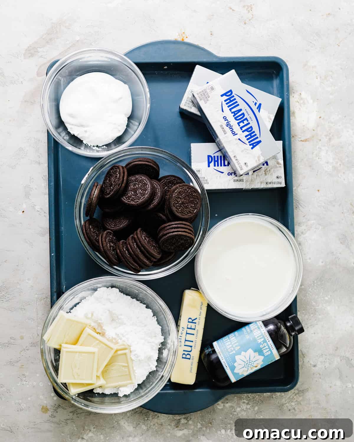 A curated display of key ingredients for making no-bake Oreo cheesecake, including blocks of cream cheese, a carton of heavy cream, a stack of Oreo cookies, white chocolate squares, vanilla extract, and powdered sugar, all arranged on a rustic wooden surface.