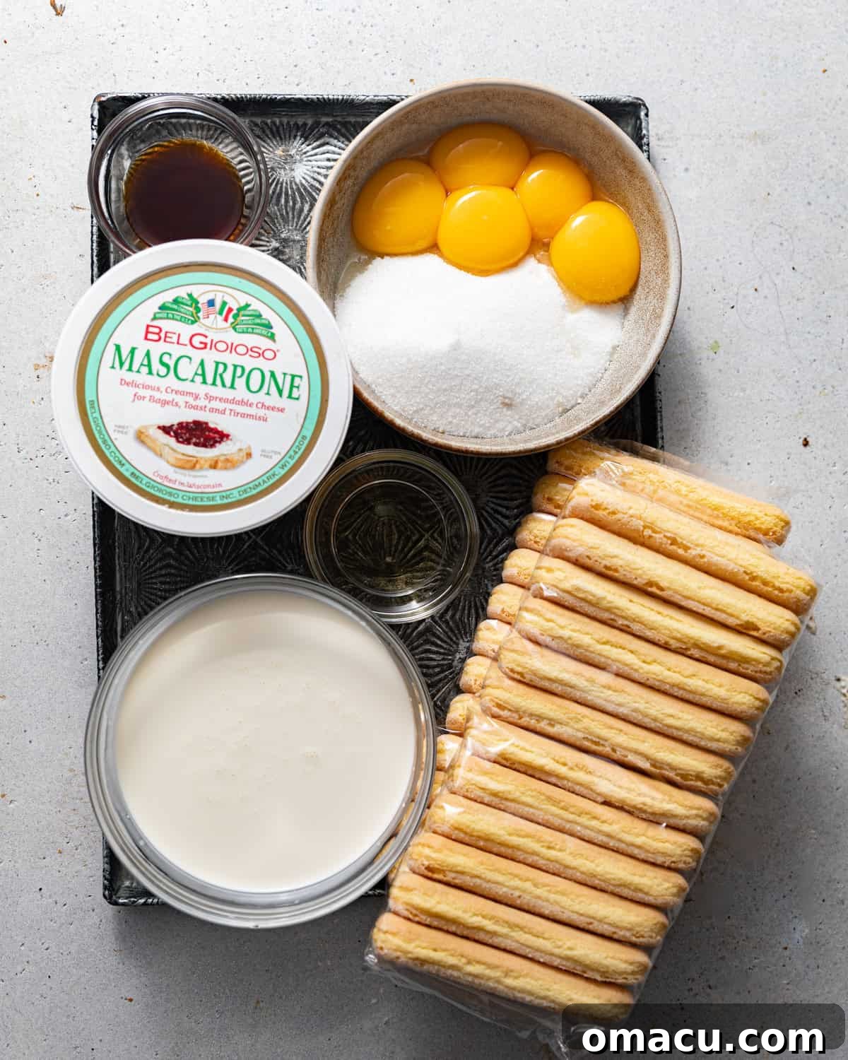 A flat lay of all essential ingredients for making authentic Italian tiramisu: egg yolks, ladyfingers, granulated sugar, vanilla extract, espresso powder, creamy mascarpone cheese, cold heavy cream, and rich cocoa powder.