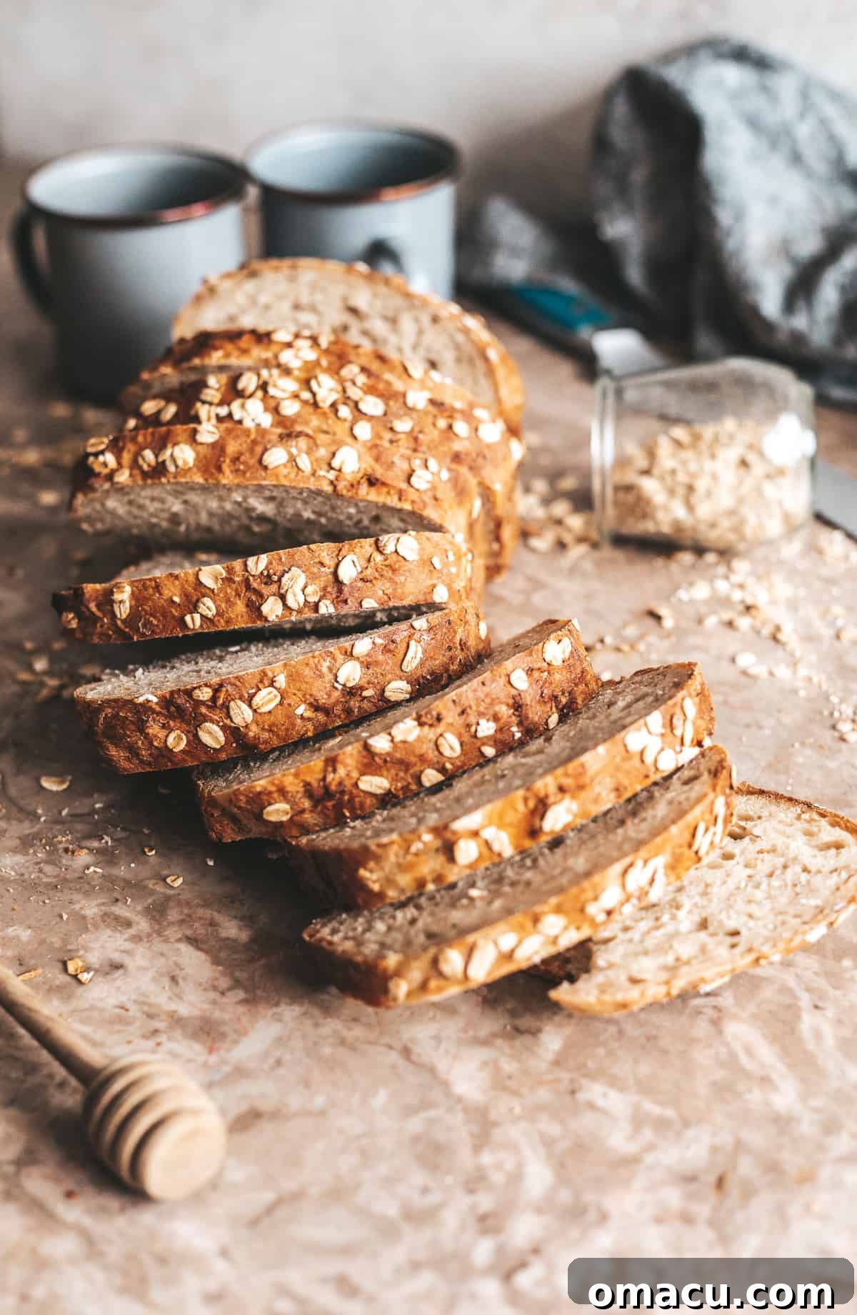 Sliced honey oat bread on a table with oats and coffee mugs, creating a cozy breakfast scene.