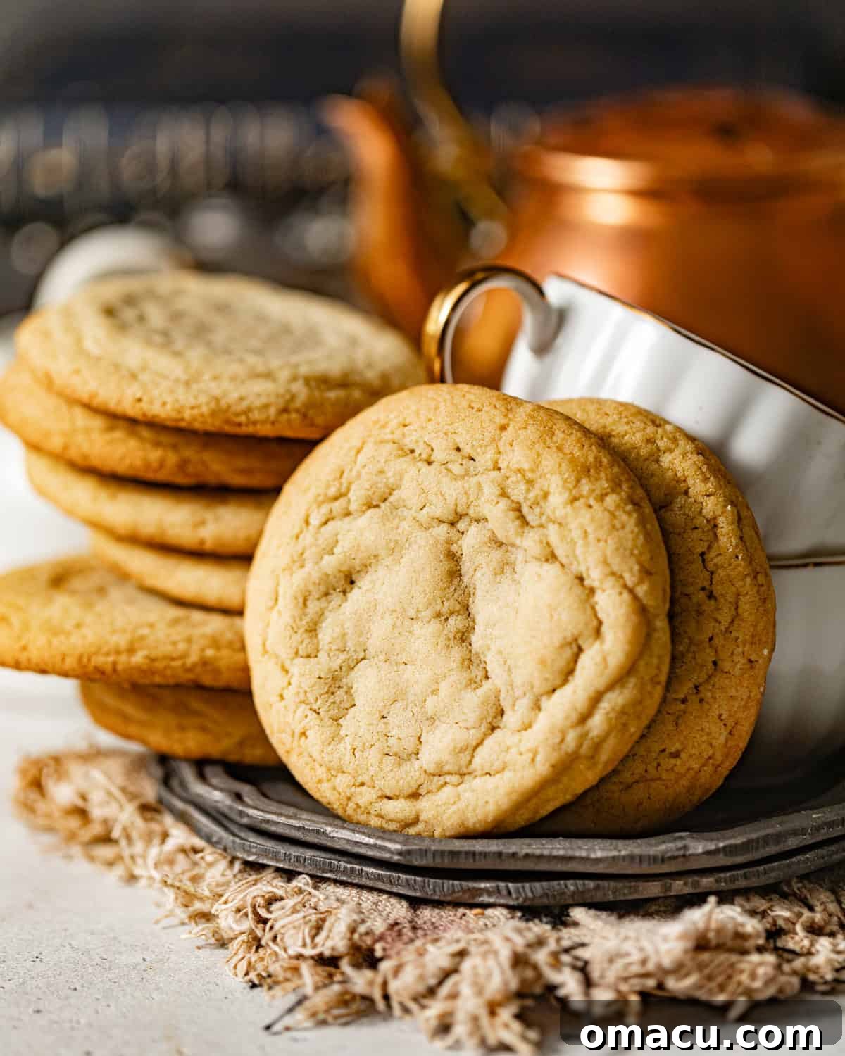 A delightful arrangement of golden-brown chocolate chipless cookies on a white plate, accompanied by two elegant teacups, suggesting a perfect pairing for a warm beverage.