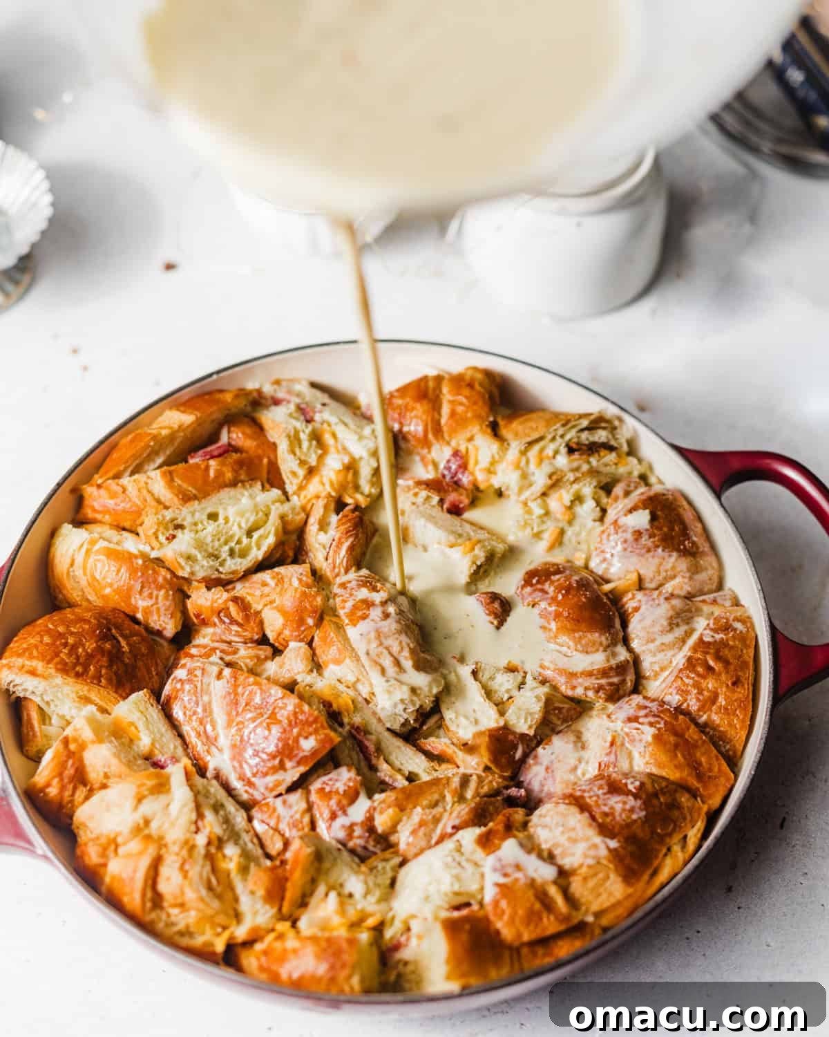 Pouring the creamy egg and milk custard mixture over the arranged croissant pieces in a casserole dish.