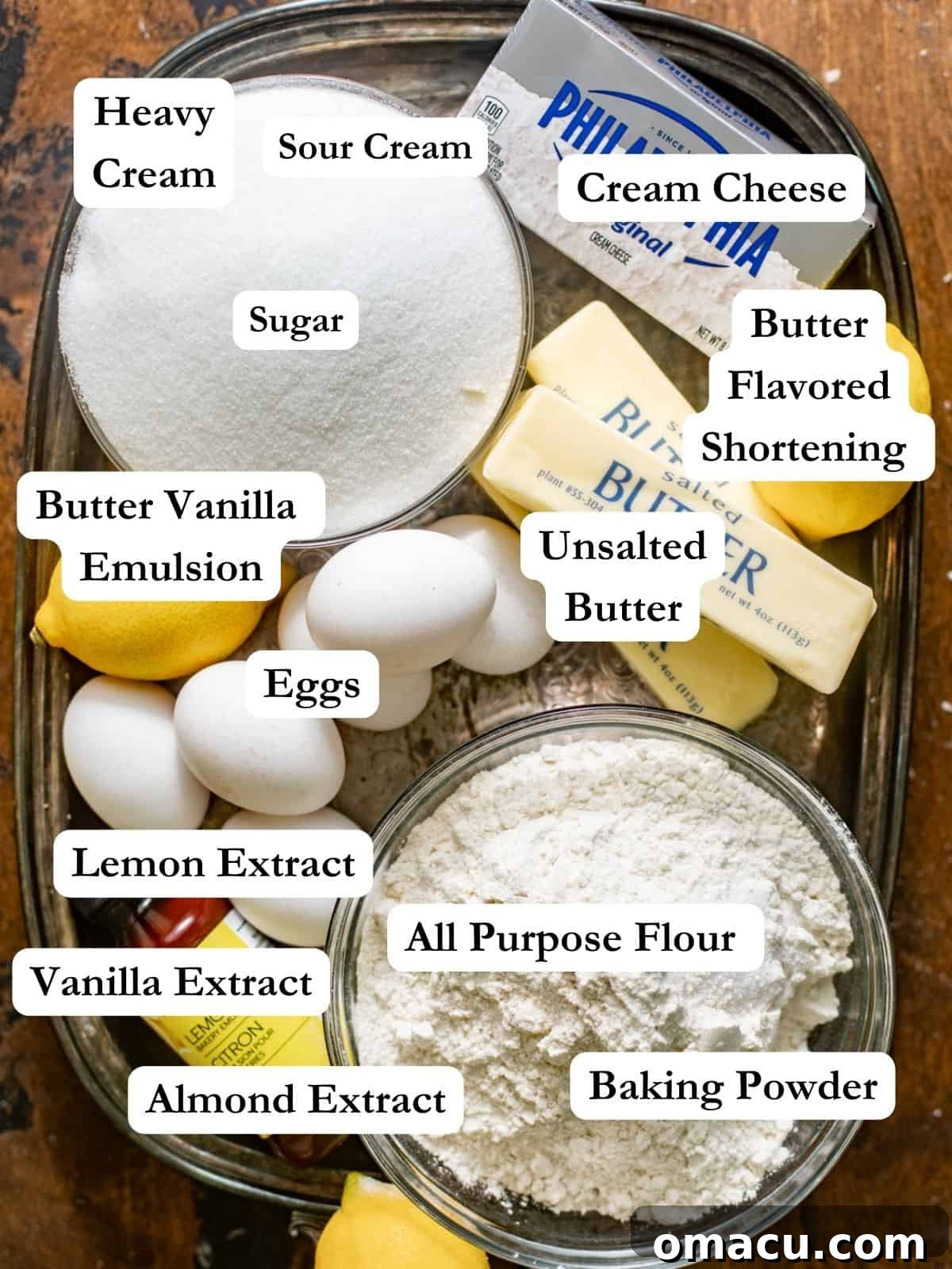 An array of cream cheese pound cake ingredients neatly arranged on a serving tray: butter, sugar, shortening, cream cheese, eggs, vanilla extract, lemon extract, almond extract, butter vanilla emulsion, heavy cream, sour cream, all-purpose flour, kosher salt, and baking powder.