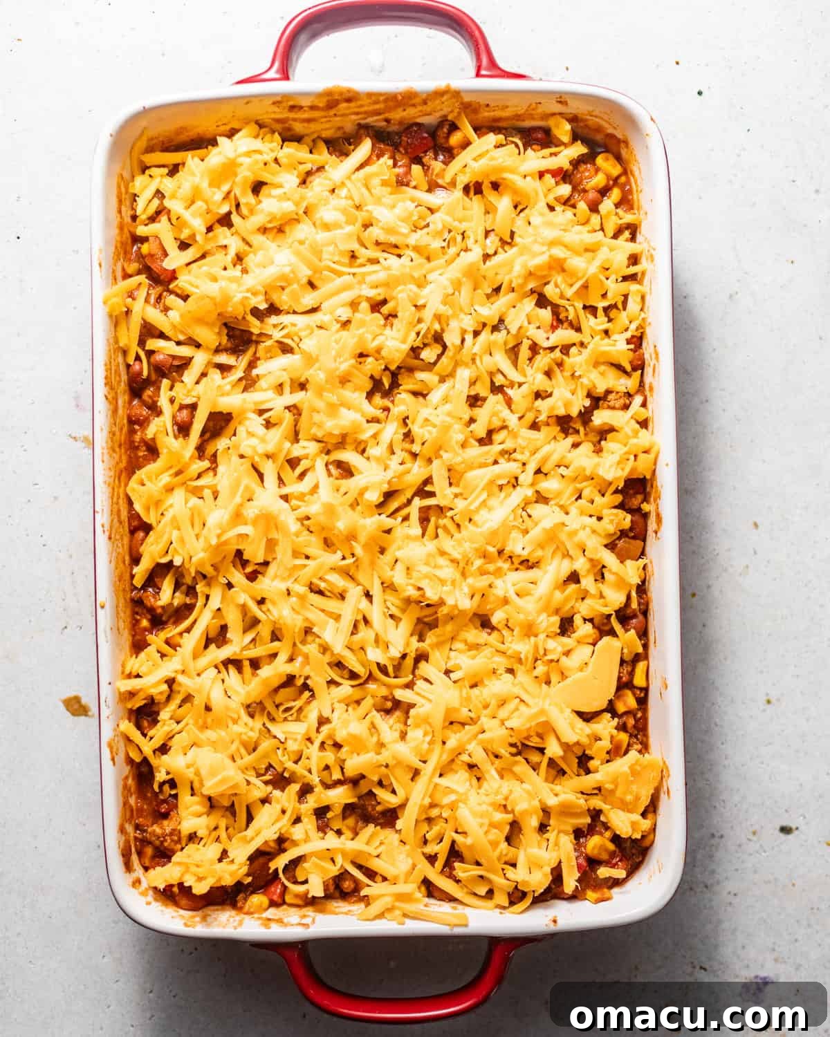 The cowboy casserole base in a baking pan, generously topped with shredded cheddar cheese, ready for the cornbread layer.
